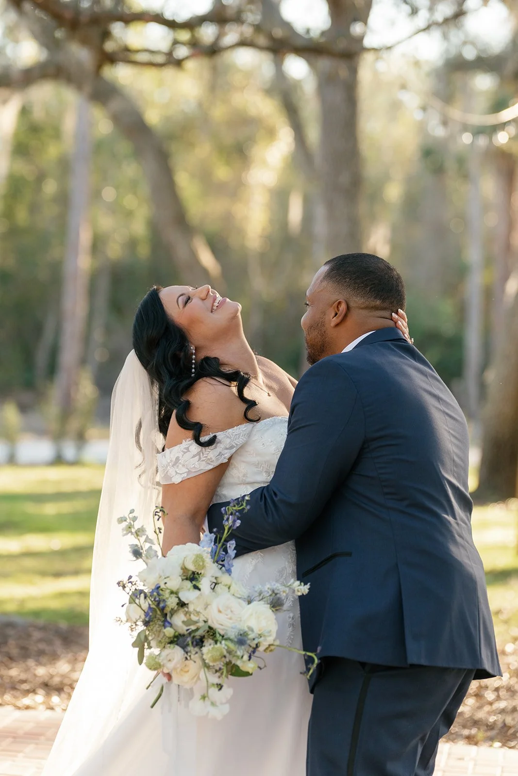 A bride and groom embrace outdoors during their wedding, with the bride smiling and holding a bouquet of white and purple flowers, and the groom gently holding her face, surrounded by trees and natural sunlight.
