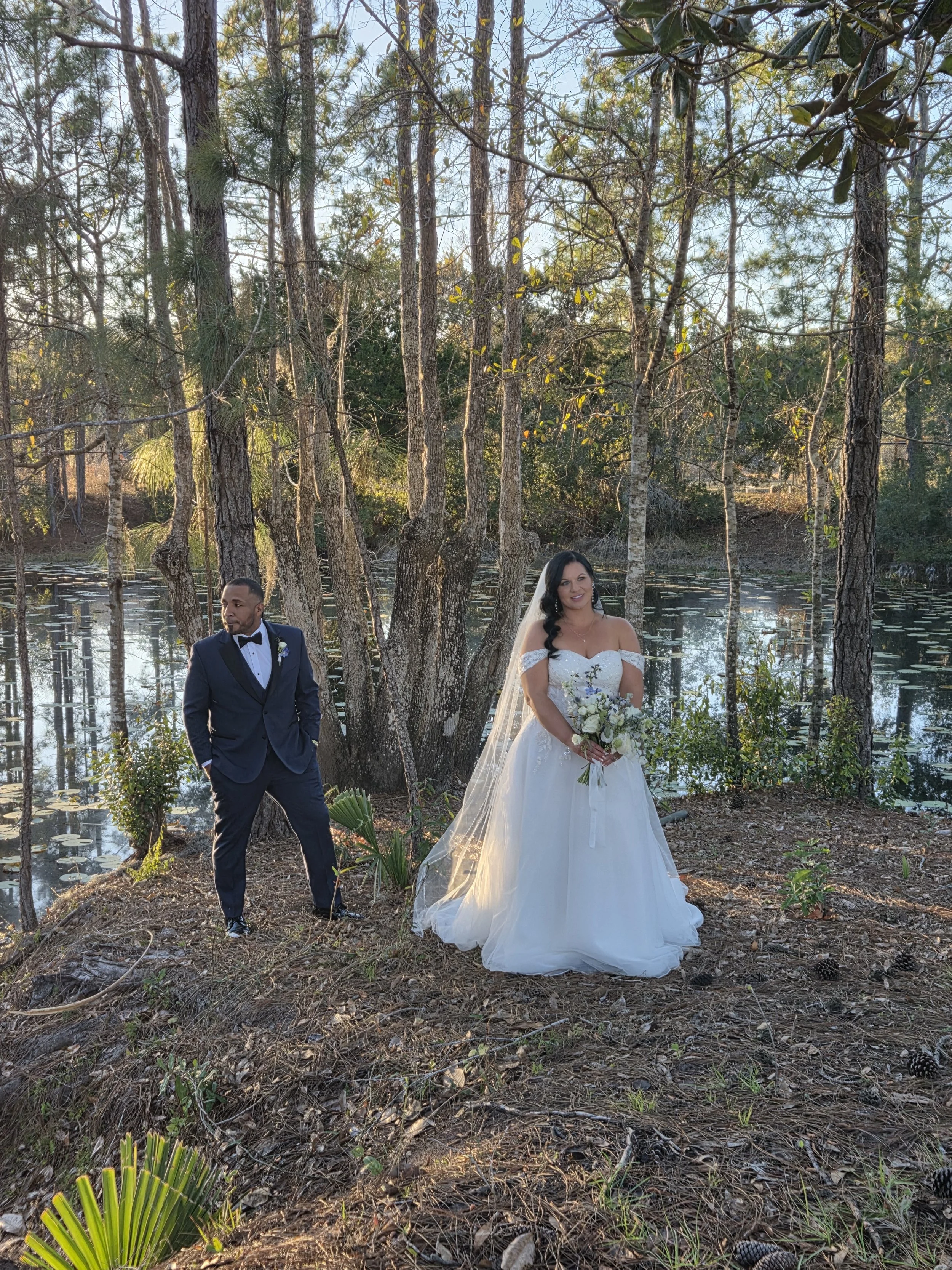 A bride in a white wedding dress holding a bouquet standing near a lake with tall trees around, and a groom in a dark suit standing nearby in a wooded outdoor setting.