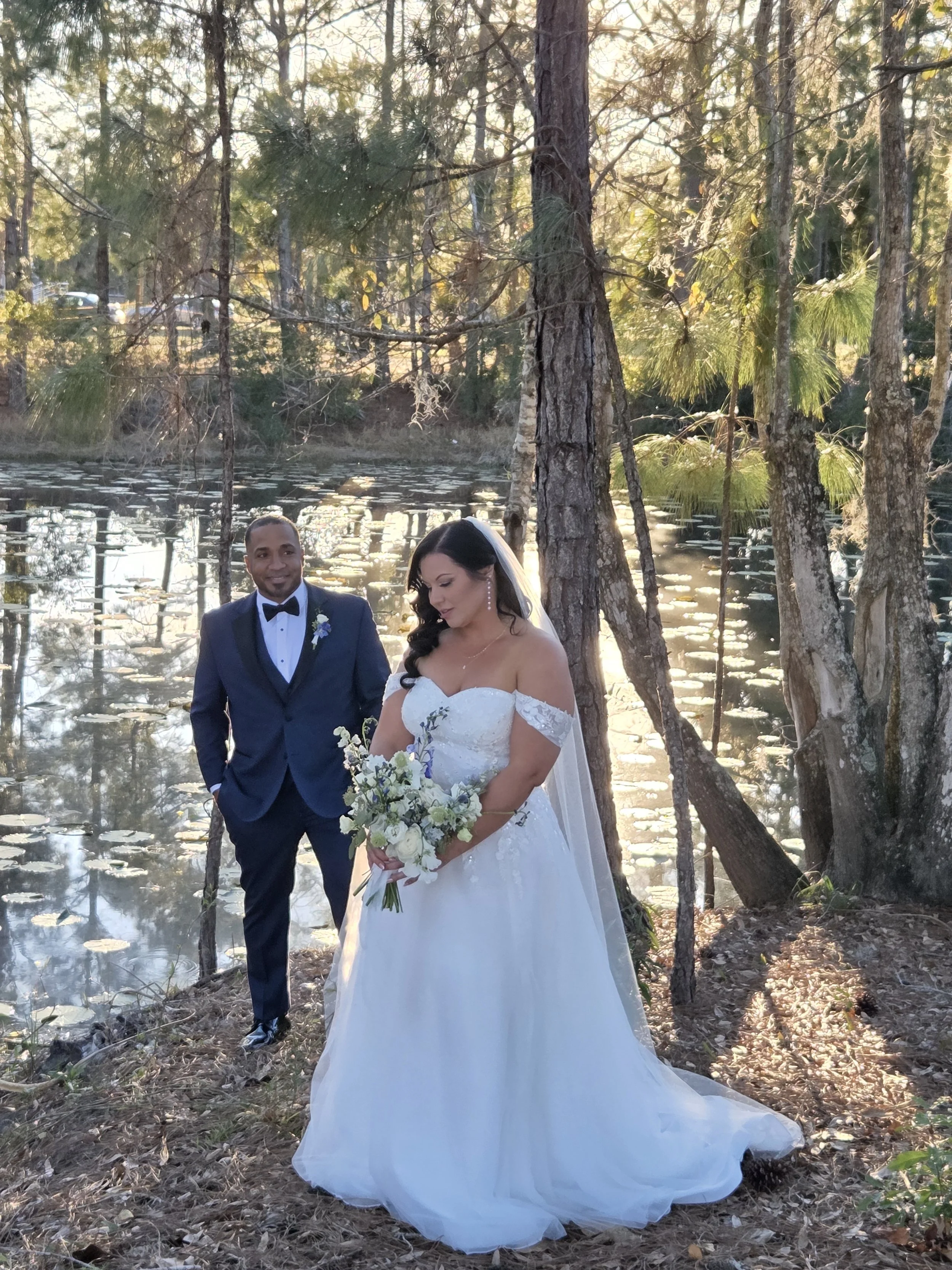 A bride and groom standing by a lake surrounded by trees during a wedding ceremony.
