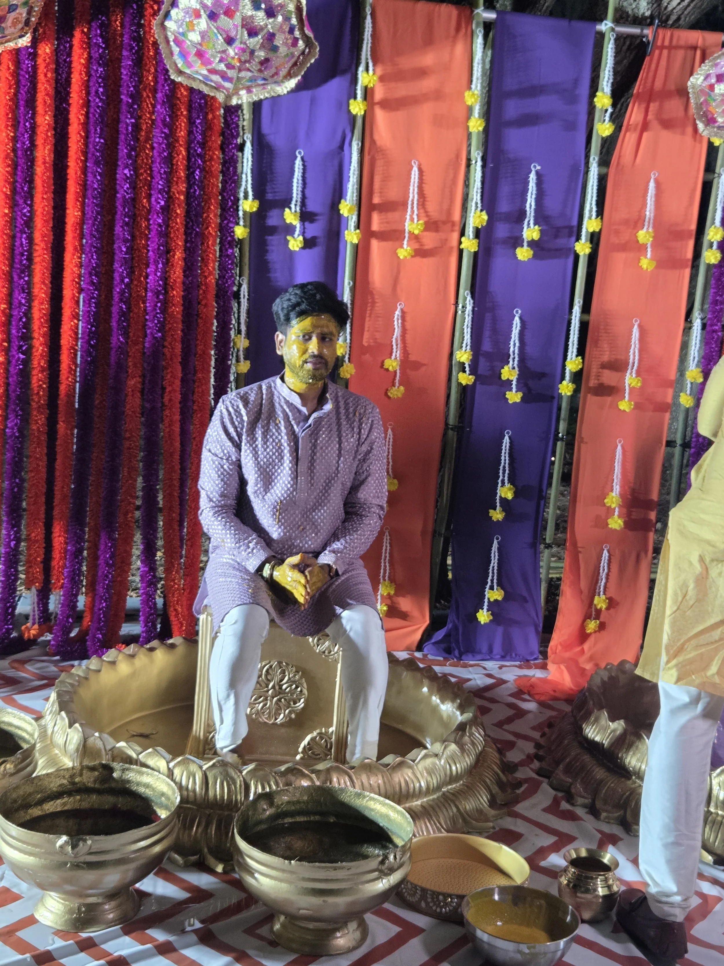 A man seated on a decorated stage during a traditional Indian ceremony, with a yellow turmeric paste on his face. The stage is adorned with purple and orange drapes, marigold flower garlands, and a large decorative canopy above. In front of him are b