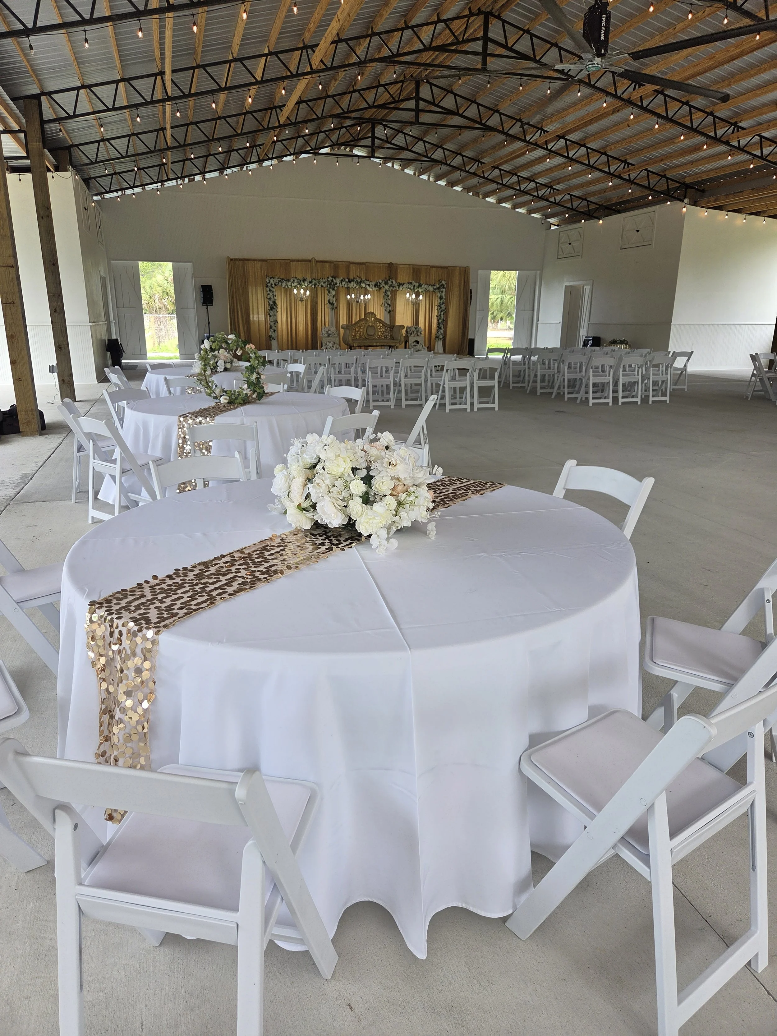 Indoor wedding venue with decorated table and chairs, floral arrangements, and a stage with a golden backdrop.