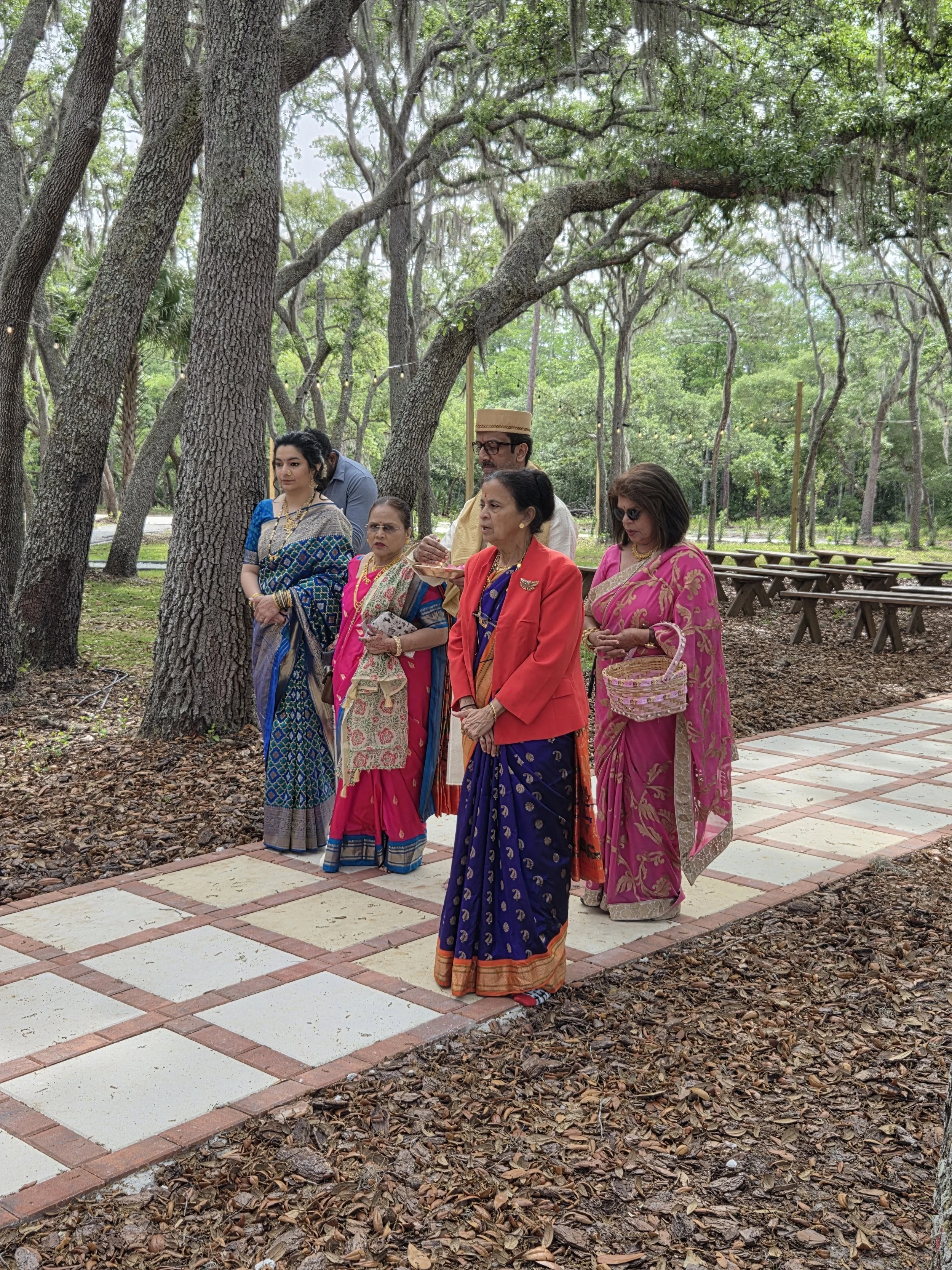 A group of six people dressed in colorful traditional South Asian clothing, standing on a paved path in a wooded outdoor area with tall trees and string lights.