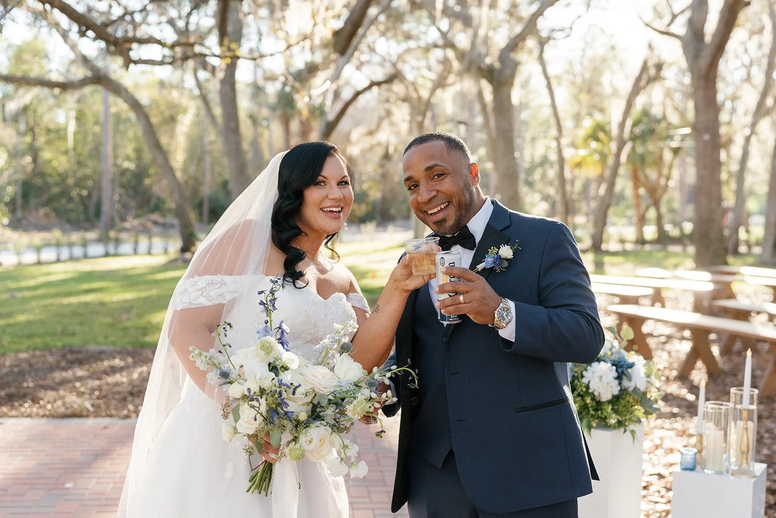 A bride and groom celebrating outdoors, holding drinks and smiling.