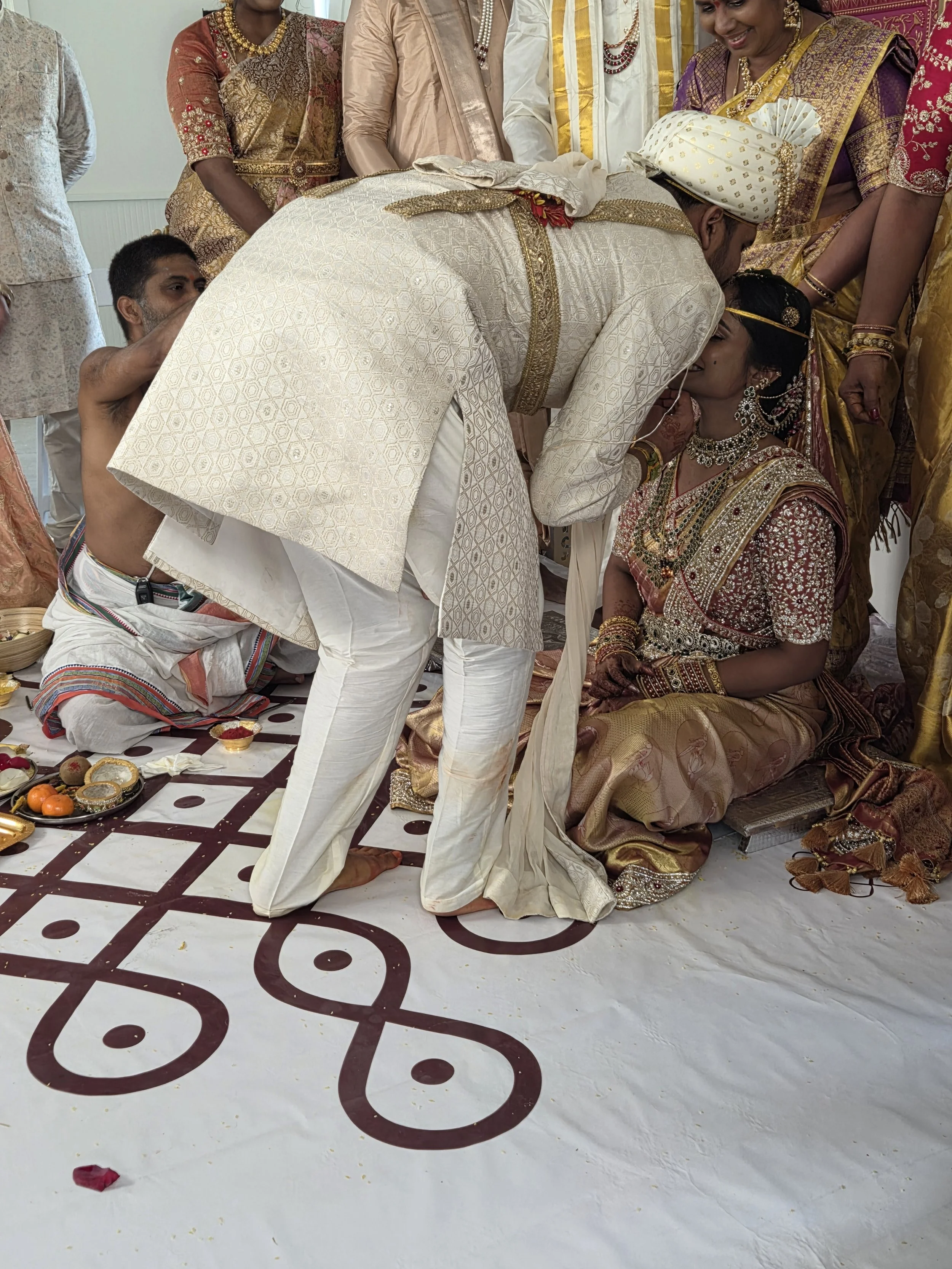 A traditional Indian wedding ceremony with the bride and groom exchanging vows. The groom, dressed in white and gold, leans forward to kiss the bride, who is wearing ornate jewelry and a richly decorated sari. Several women in colorful sarees and jew