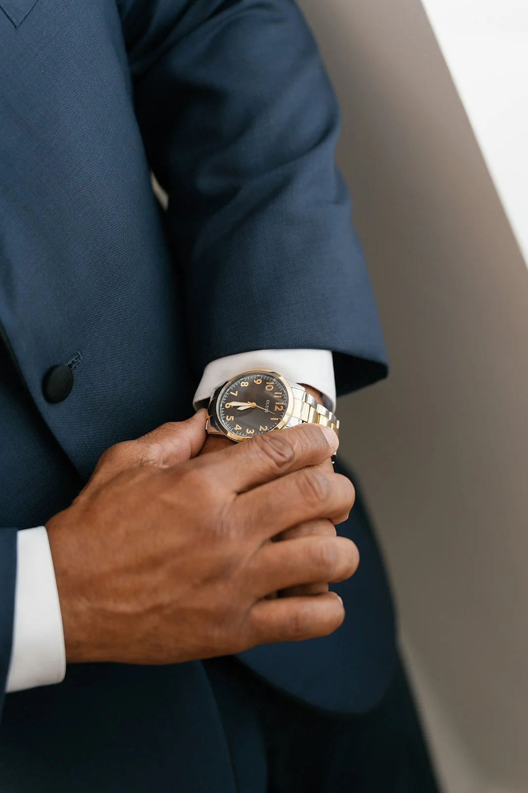 Close-up of a person's wrist showing a gold and silver watch, with the person dressed in a dark blue suit and white shirt, sitting with their hand resting on their lap.