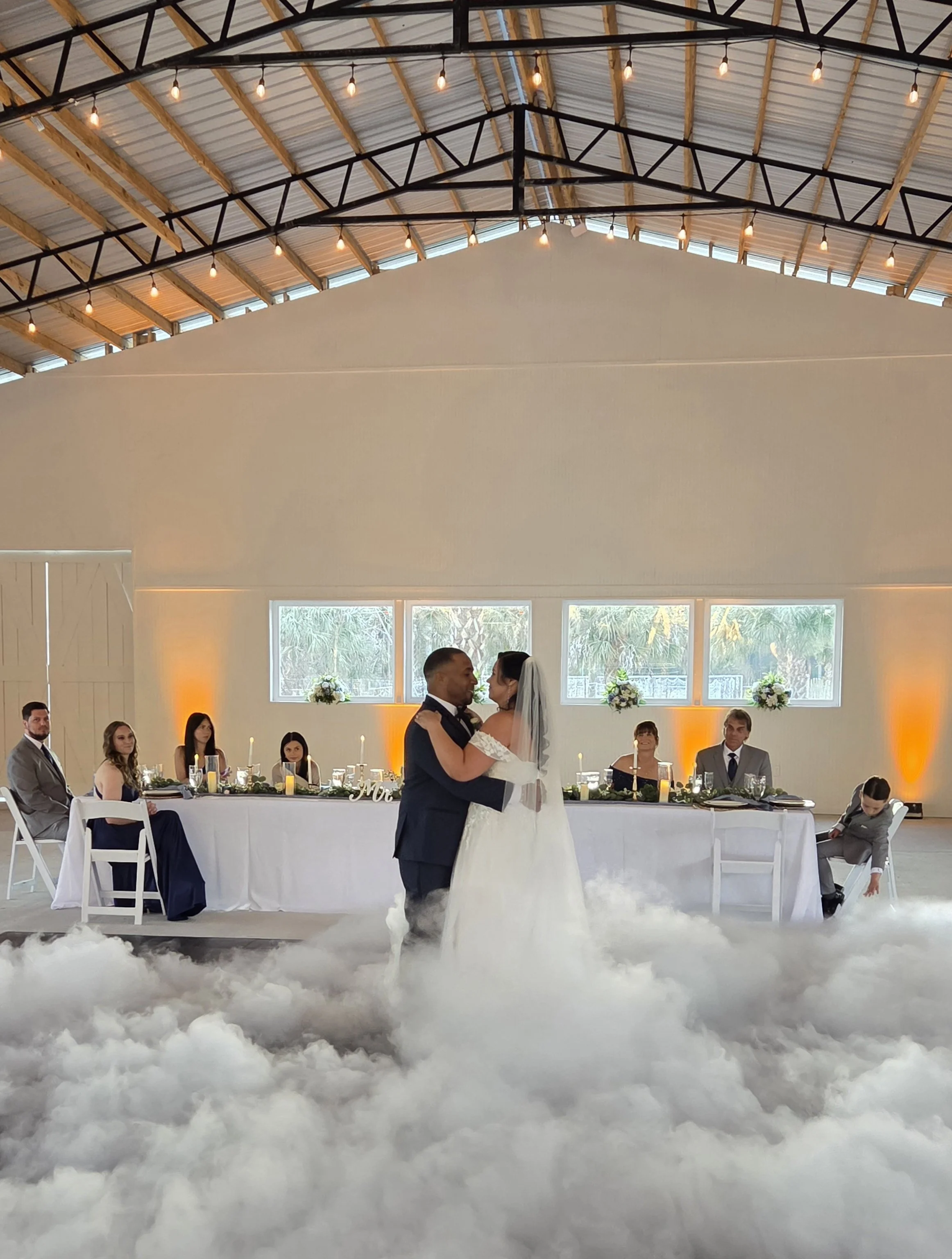 A bride and groom dancing at their wedding reception inside a large venue with high vaulted ceiling and exposed beams. The guests are sitting at a long table decorated with candles and floral arrangements, with a cloud-like floor covering beneath the