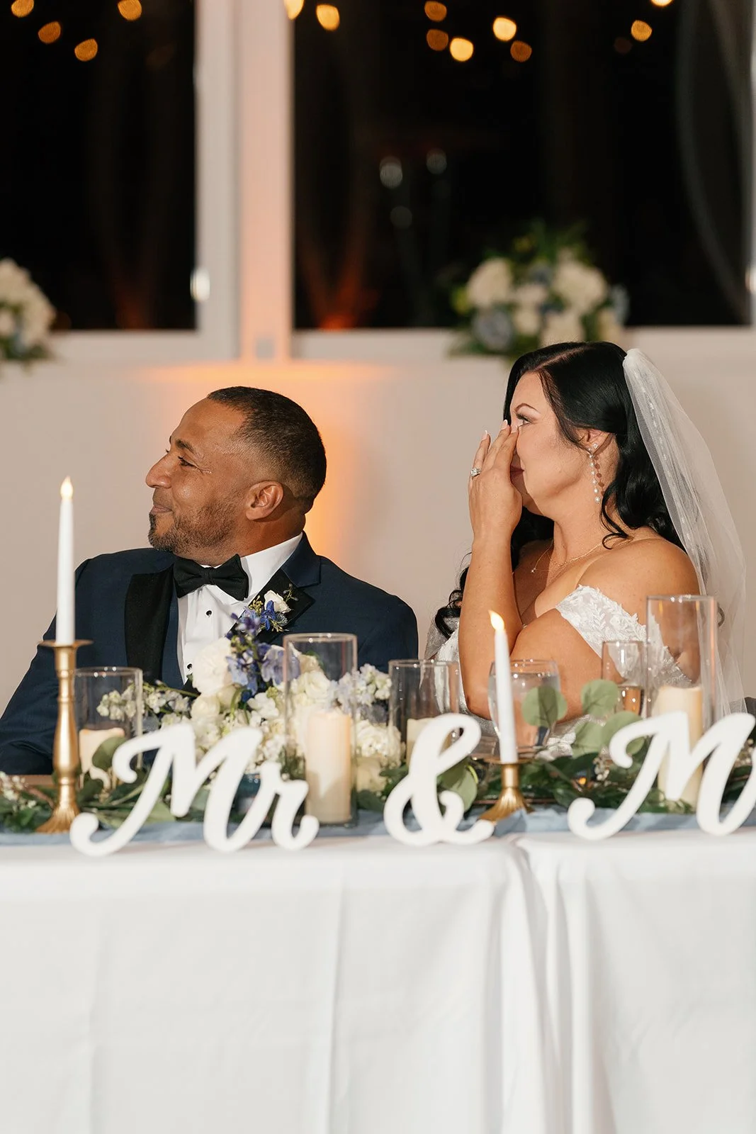 Bride and groom sitting at a wedding reception table, with the bride covering her face with her hand and the groom smiling. The table has candles, flowers, and a sign that reads 'Mr & M'.