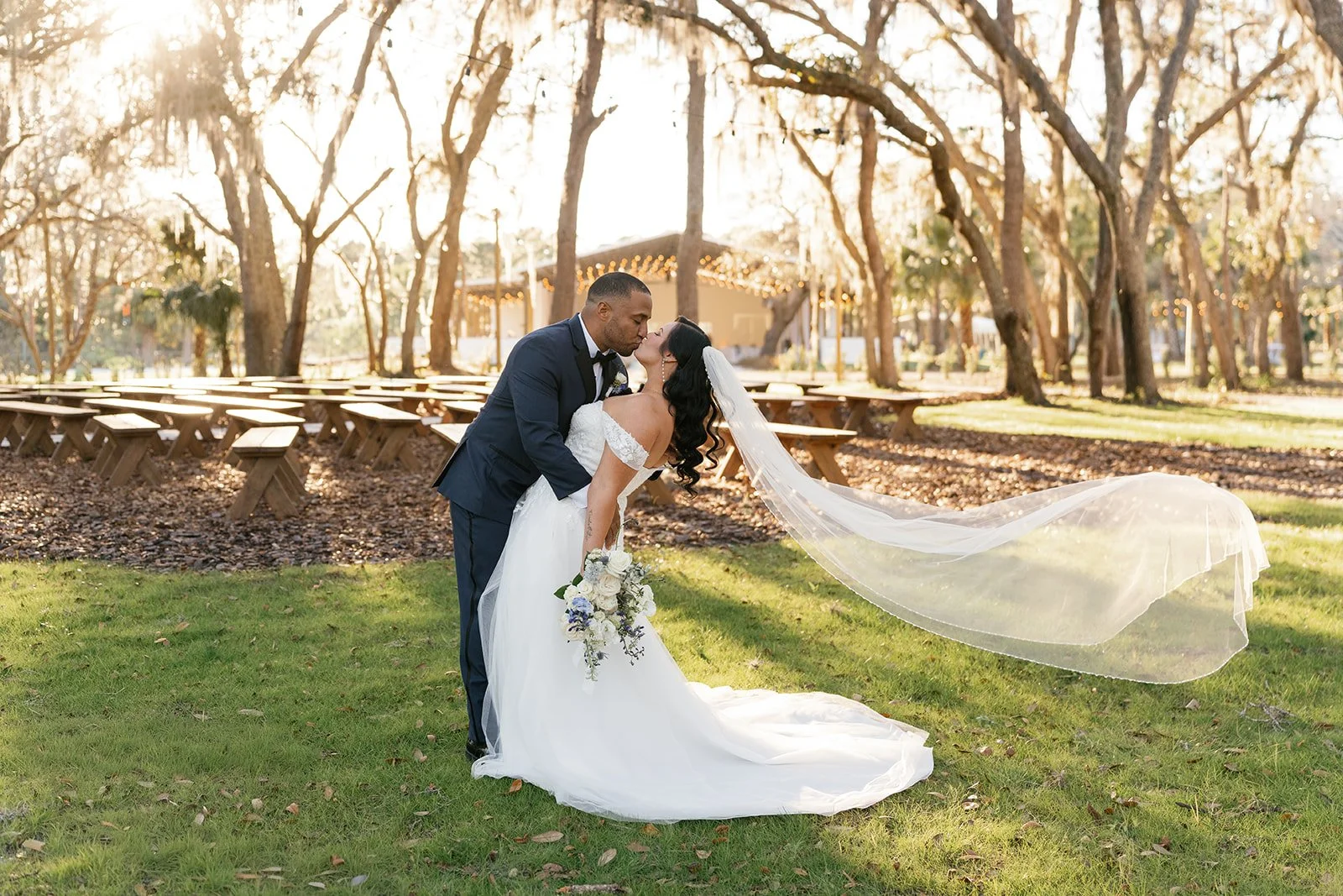 A newlywed couple sharing a romantic kiss in a park at sunset, with the groom dipping the bride. The bride is wearing a white wedding dress with a long veil and holding a bouquet. The groom is in a navy suit with a bow tie. Leafy trees and wooden ben