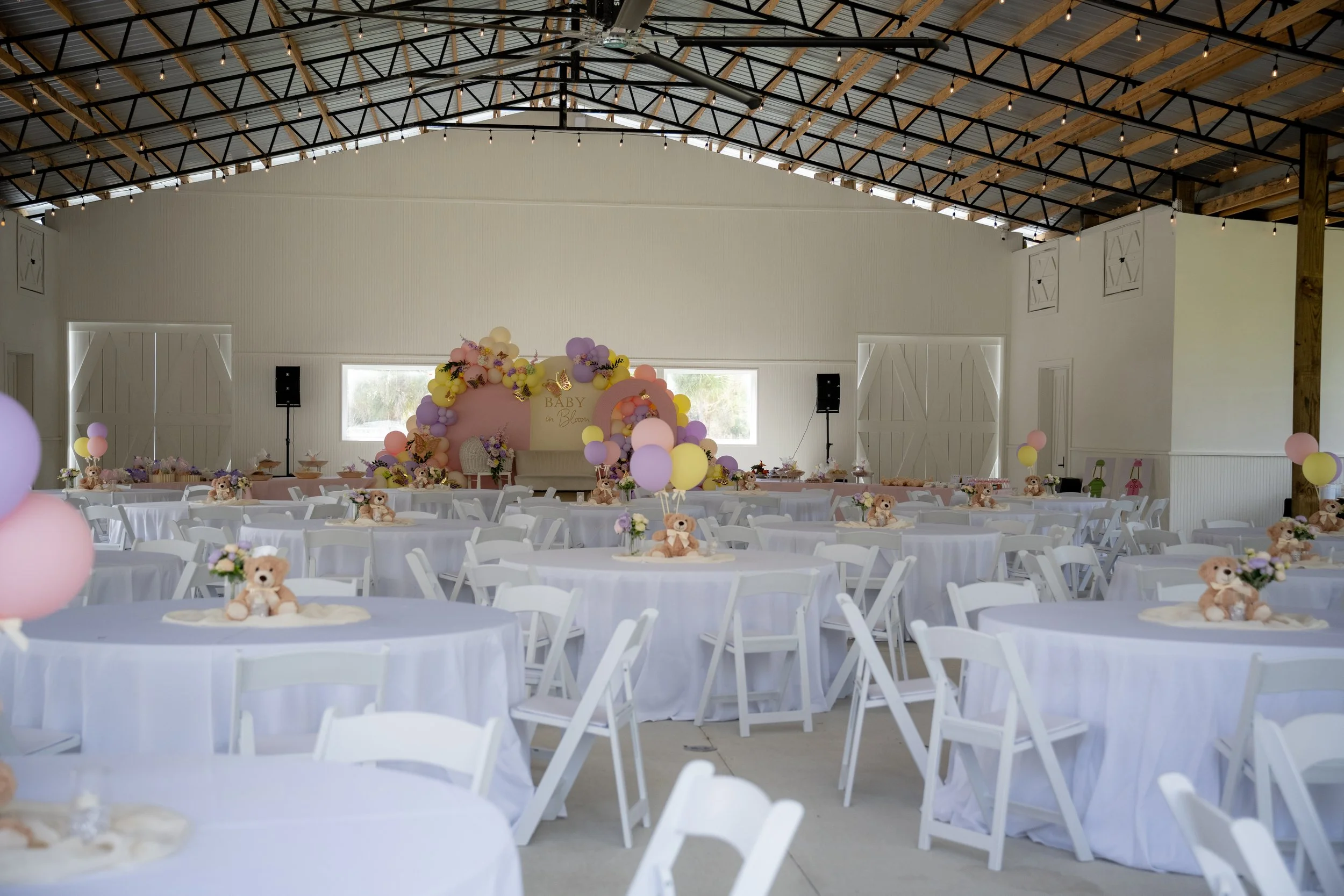 Indoor event space decorated for a baby shower with pastel-colored balloons, teddy bears, and floral arrangements. Chairs are set around round tables covered with white tablecloths. A decorated arch with a sign reading 'Baby on Blossom' is at the fro