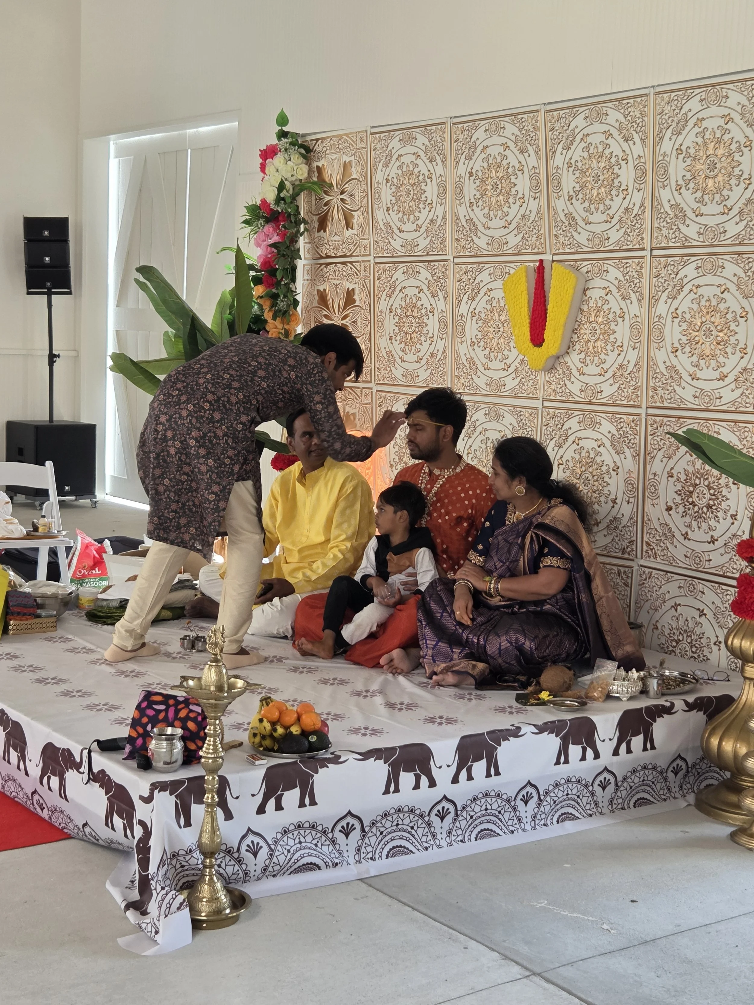 Hindu wedding ceremony setup with a decorated table, children and adults sitting and participating, traditional clothes, floral decorations, and religious items.