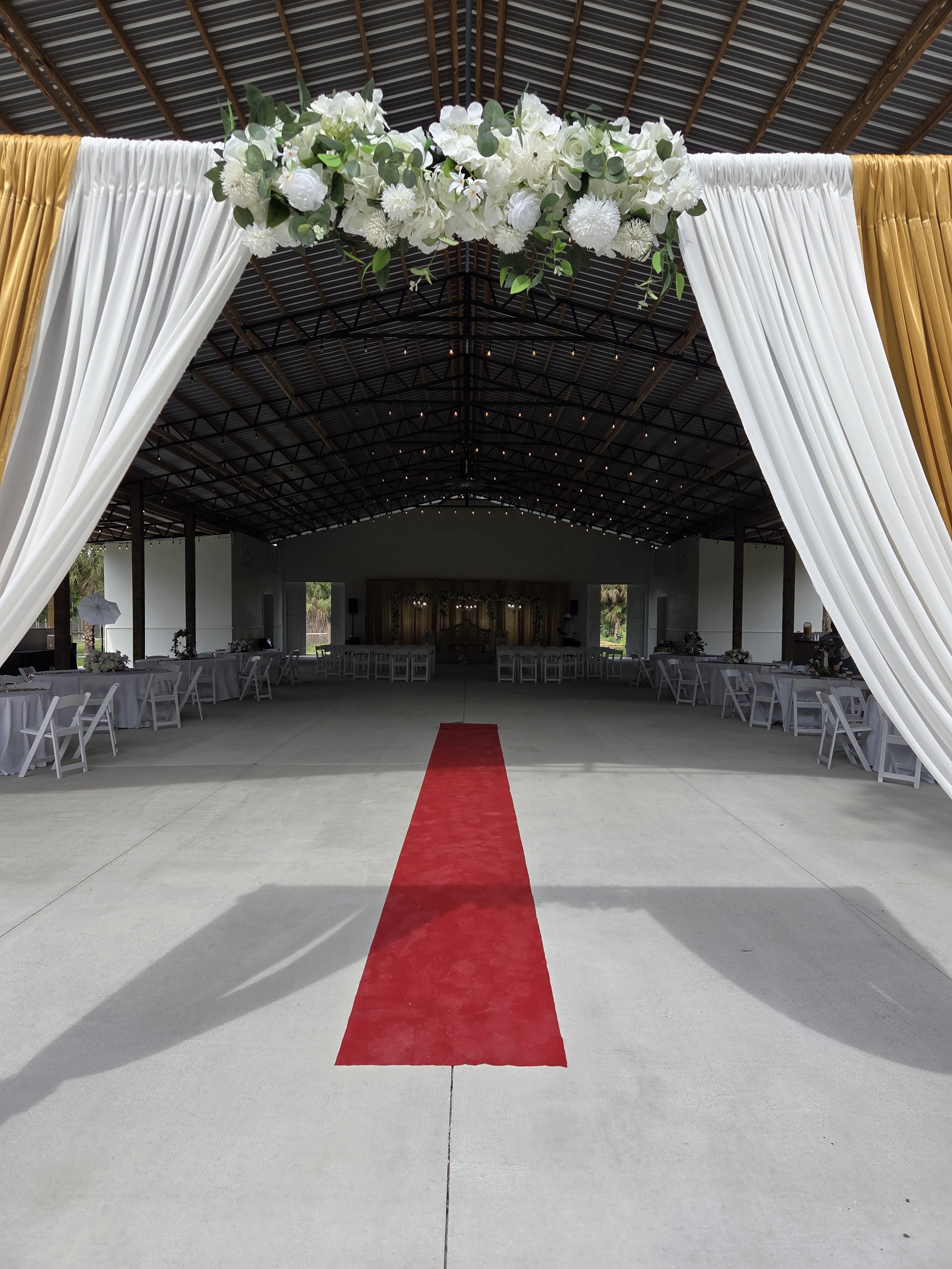 Wedding venue with a red carpet leading to the stage, decorated with white curtains, golden drapes, and a floral arrangement overhead.