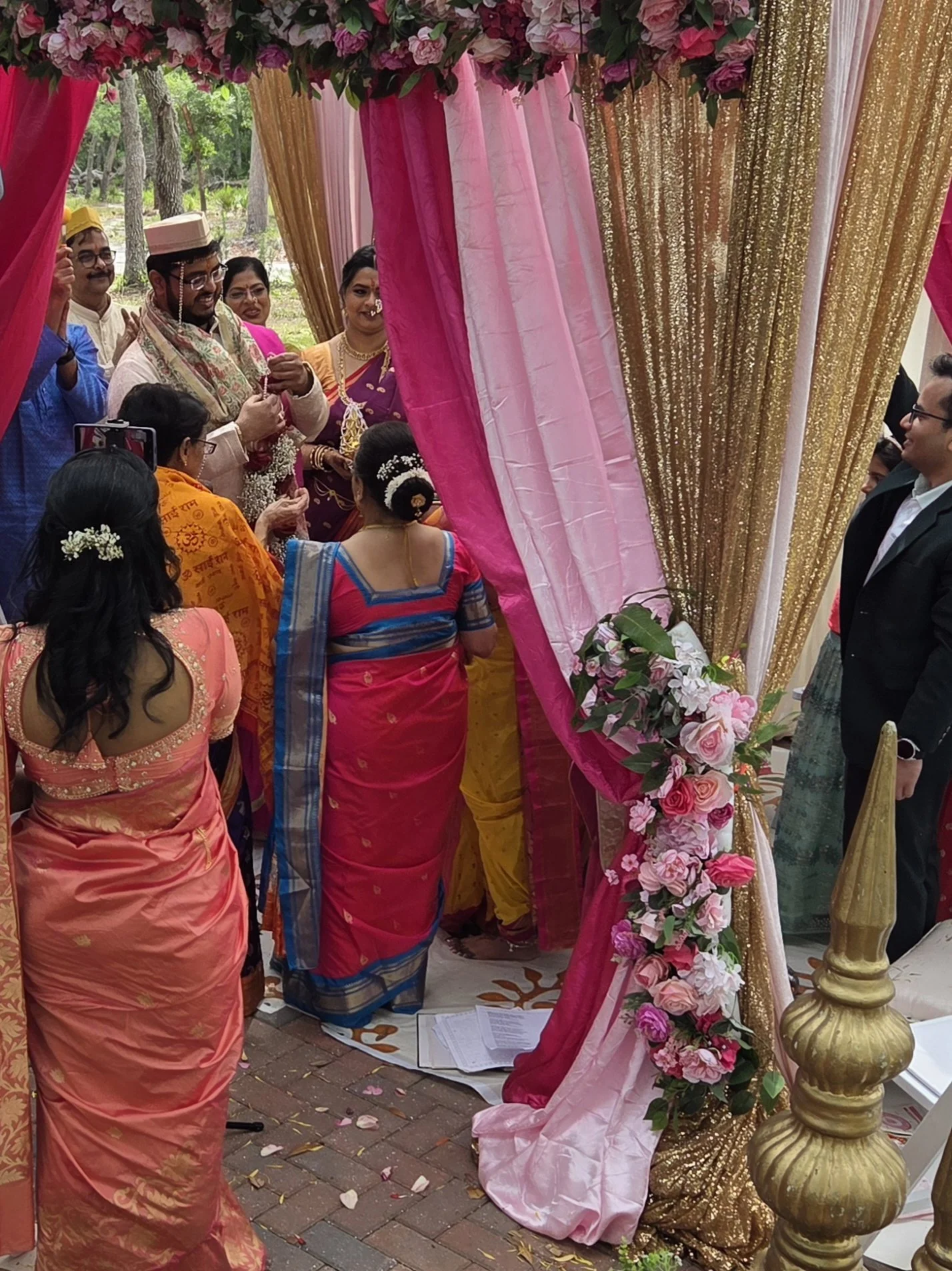 People participating in a traditional Indian wedding ceremony outdoors, under a decorated canopy with pink and gold drapes and floral arrangements.