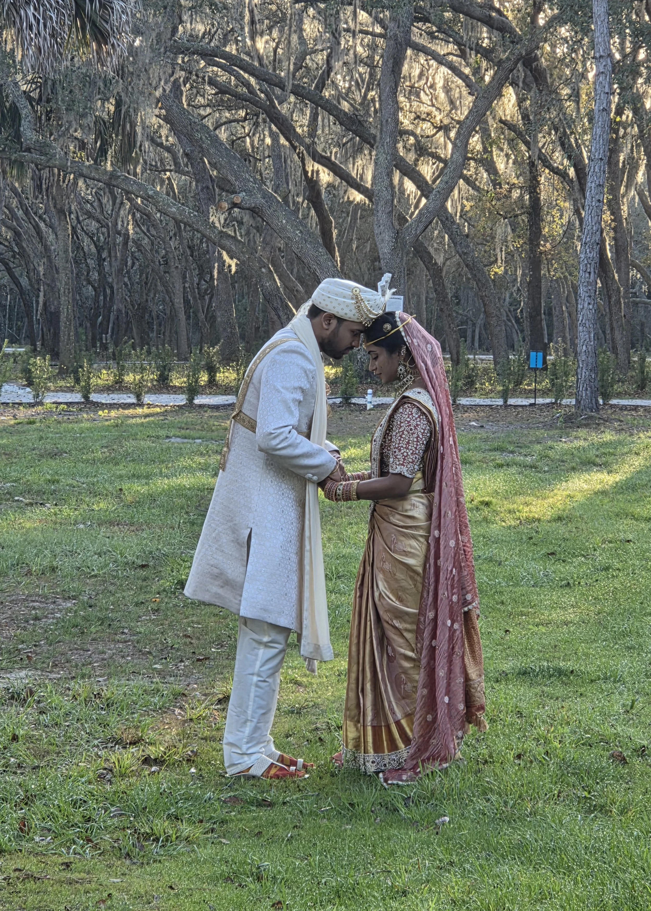 A couple dressed in traditional Indian wedding attire sharing a moment outdoors in a wooded area, with their foreheads touching and eyes closed.