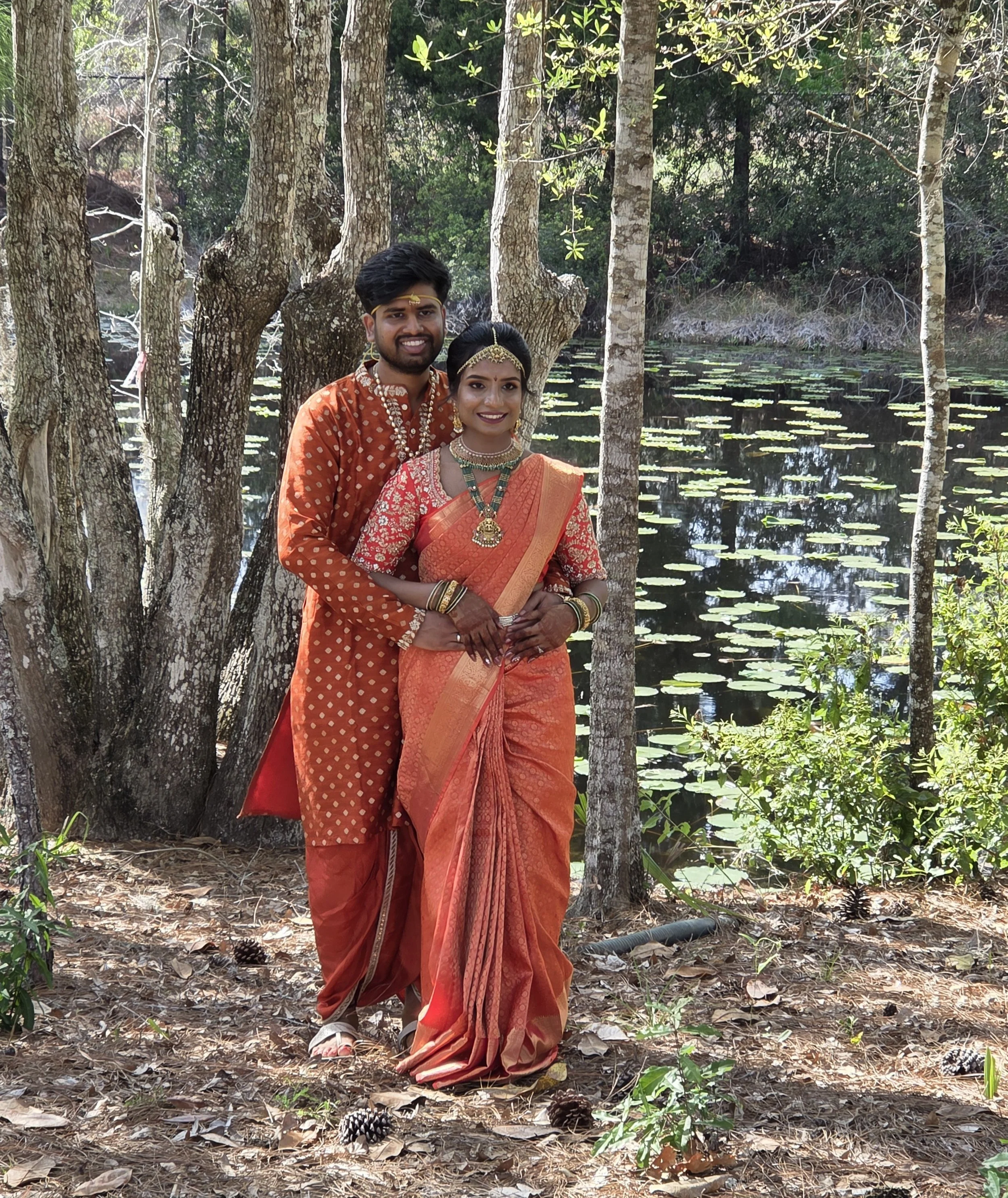 A young couple dressed in traditional South Asian attire stands outdoors near a pond surrounded by trees and foliage, smiling at the camera.