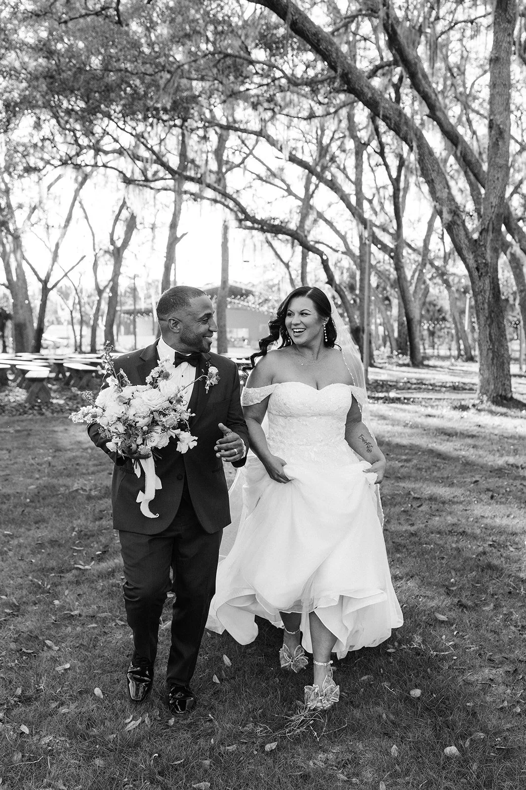 A bride and groom walking outdoors in a park, smiling, with trees in the background. The groom is holding a bouquet and wearing a tuxedo, while the bride is in a wedding gown.