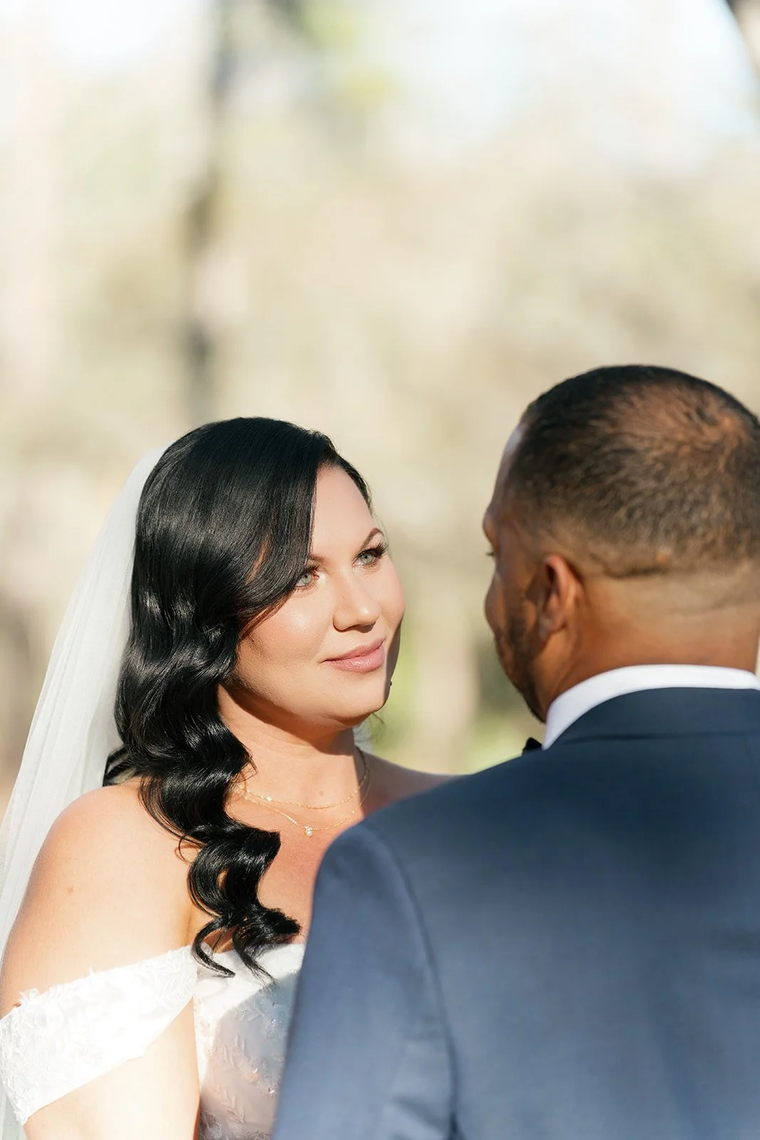 A bride with black wavy hair and a strapless wedding dress faces a groom with short hair and a dark suit during a wedding ceremony outdoors.