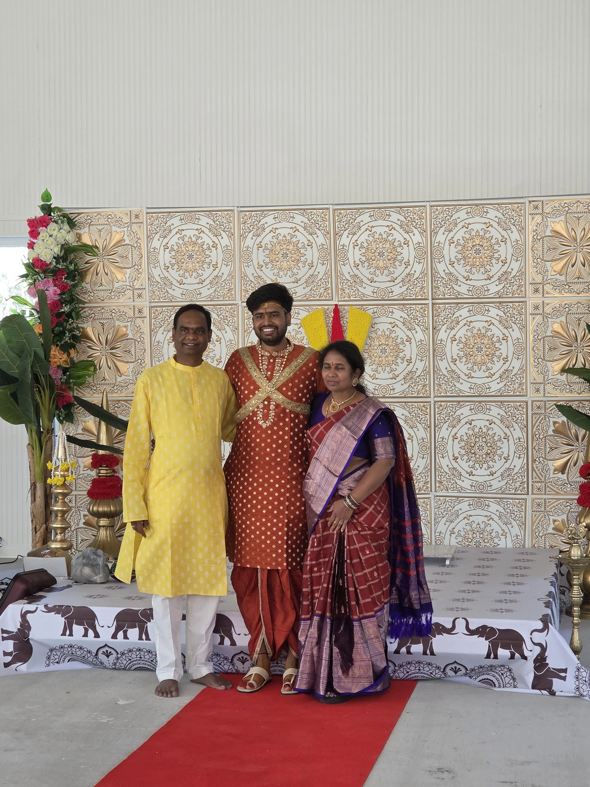Three people standing together on a stage during a celebration or ceremony. They are dressed in traditional Indian clothing. The man on the left wears a yellow kurta with white pants. The man in the middle wears a red and gold kurta with matching pan