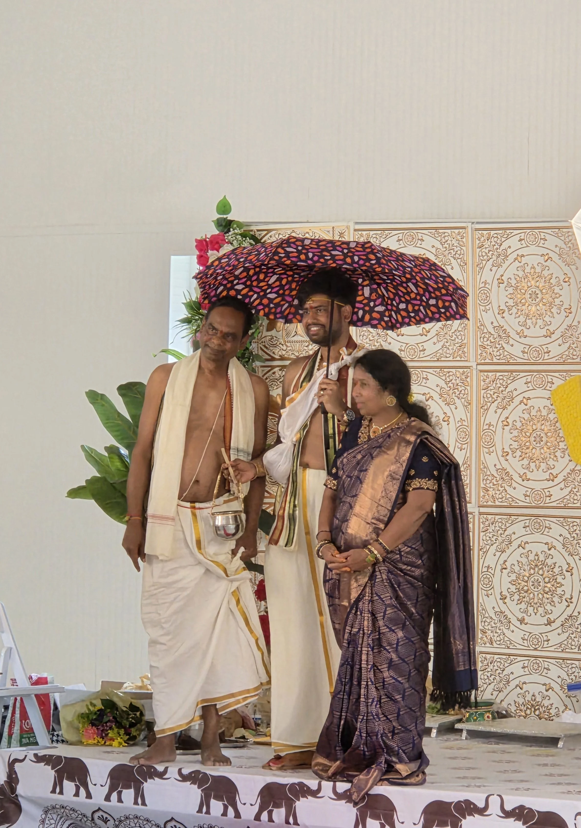 Three people participating in a traditional Indian ceremony or wedding. The man in the middle is holding a colorful umbrella over himself and two others. The woman is dressed in a sari, and the man on the left is wearing traditional attire. The backg