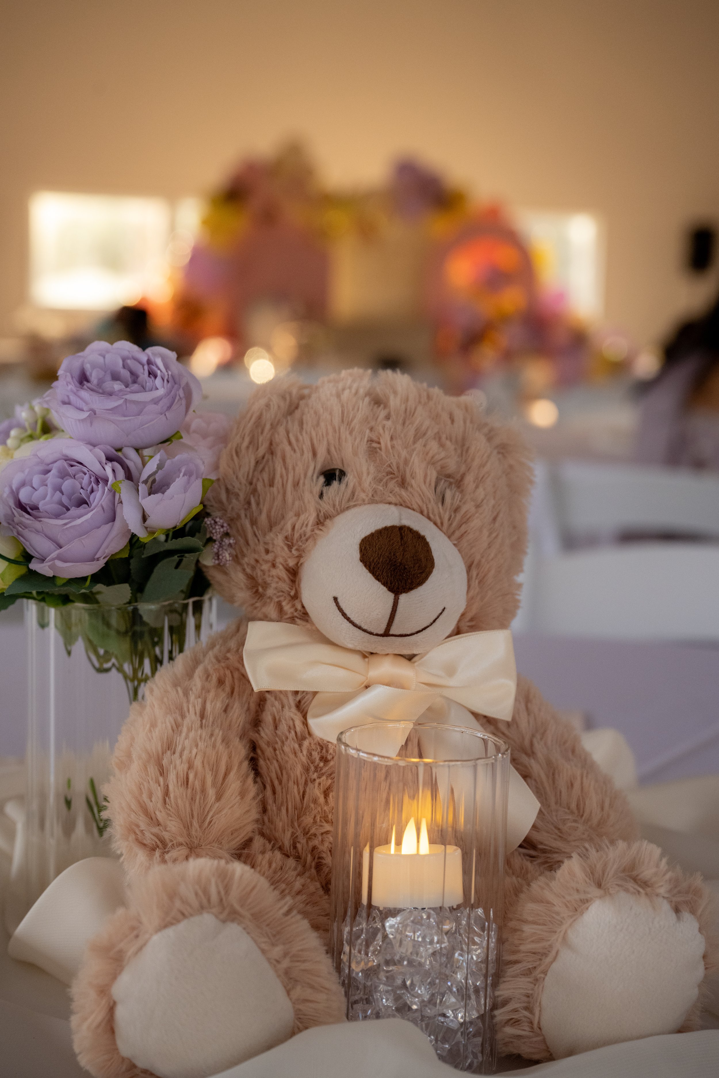 A teddy bear with a cream-colored bow tie, sitting in front of a lit candle in a glass holder, with purple flowers and a blurred colorful background.