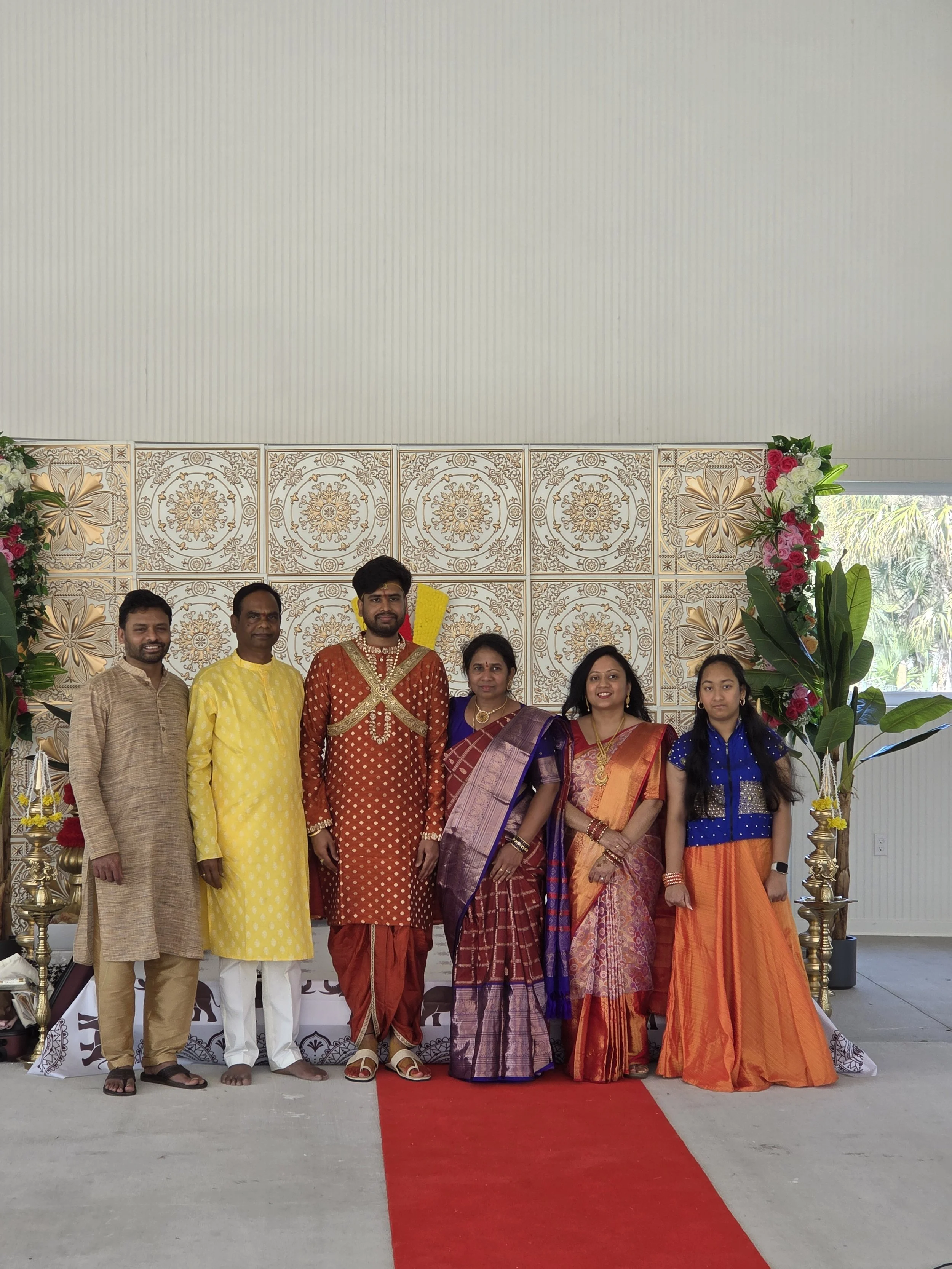 Group of seven people dressed in traditional Indian attire standing on a red carpet in front of a decorated backdrop with floral arrangements.