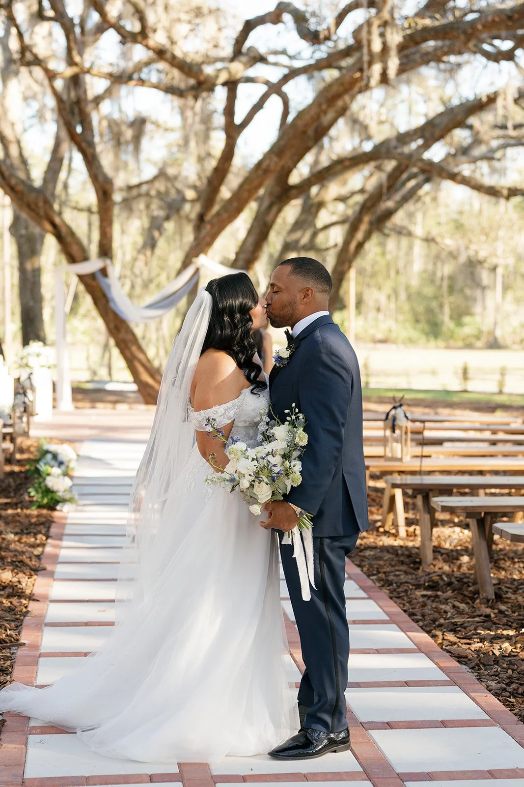 A bride and groom sharing a kiss during their outdoor wedding ceremony, with wooden benches, floral arrangements, and large trees in the background.