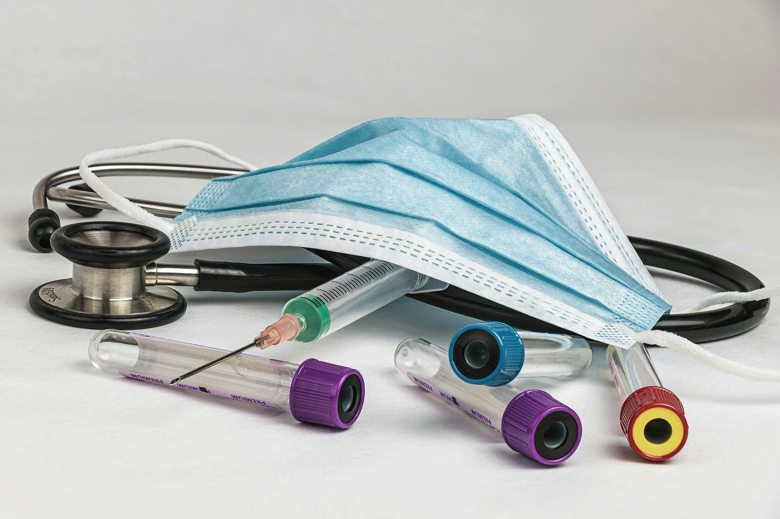 Medical items including a blue surgical mask, stethoscope, syringe, and blood collection tubes on a white surface.