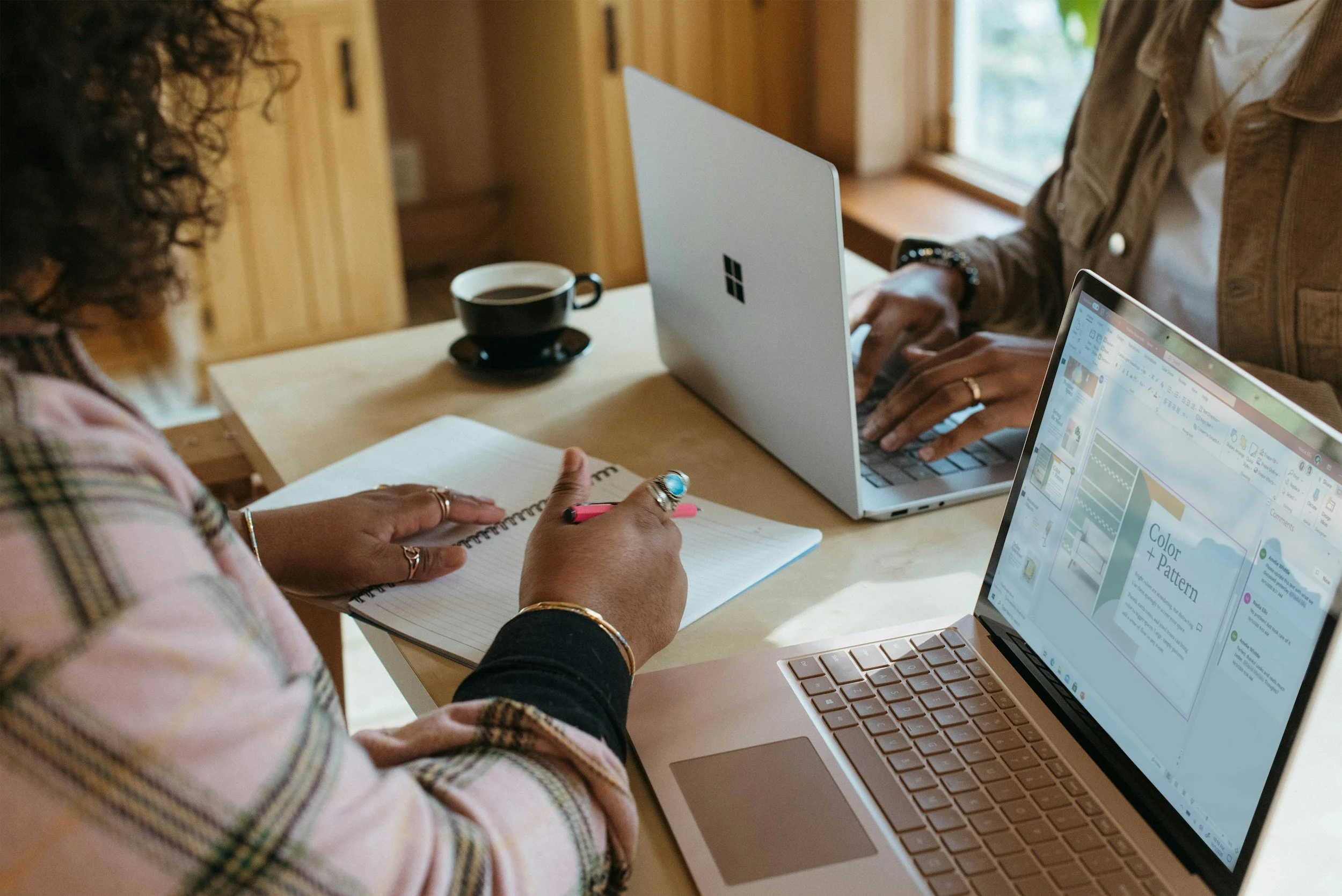 Two women working at a wooden table with laptops, notebook, and coffee, in a cozy wooden room with sunlight coming through a window.