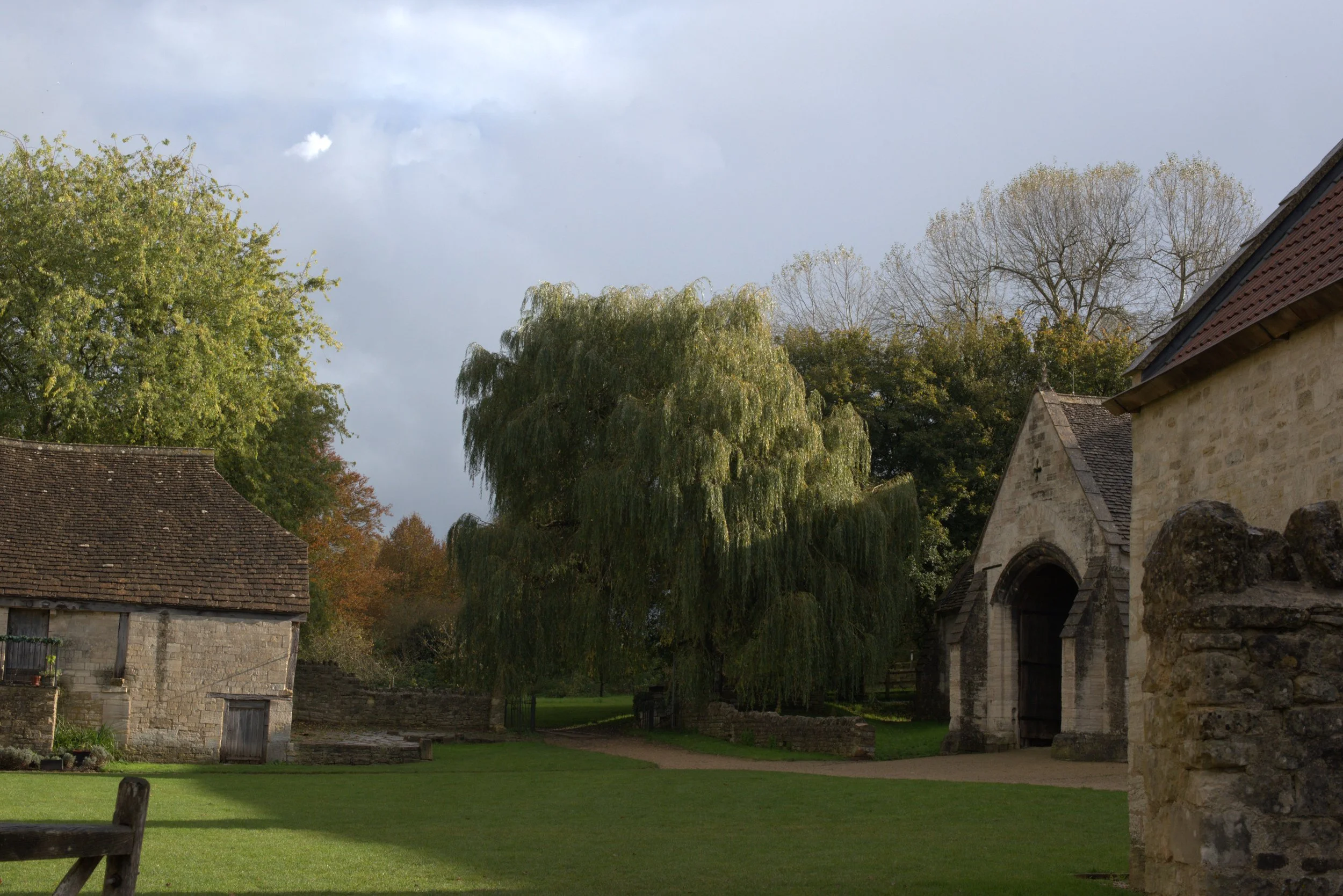 A peaceful rural scene featuring old stone buildings, a large green weeping willow tree, and a well-kept grassy yard under a partly cloudy sky.