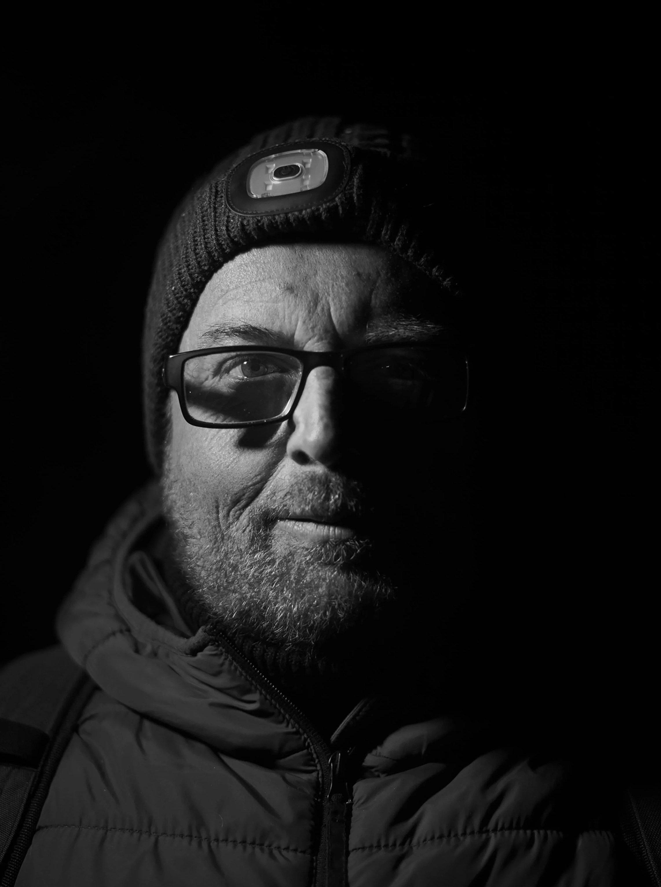 A man wearing glasses, a beanie with a headlamp, and a puffy jacket, illuminated from one side in a dark environment.