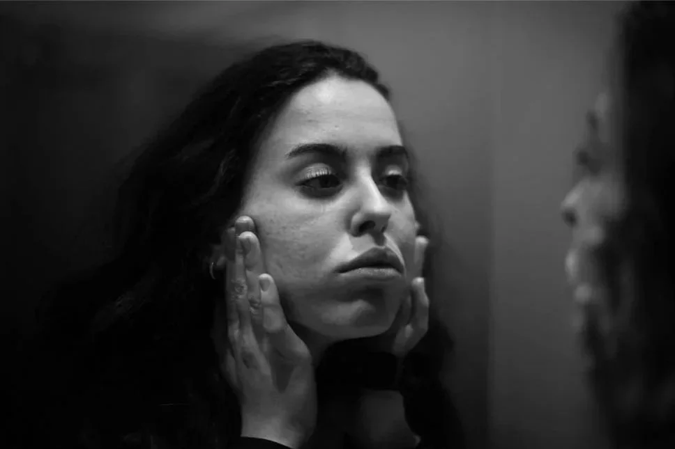 Woman looking at her reflection in a mirror, touching her face with both hands, in black and white.