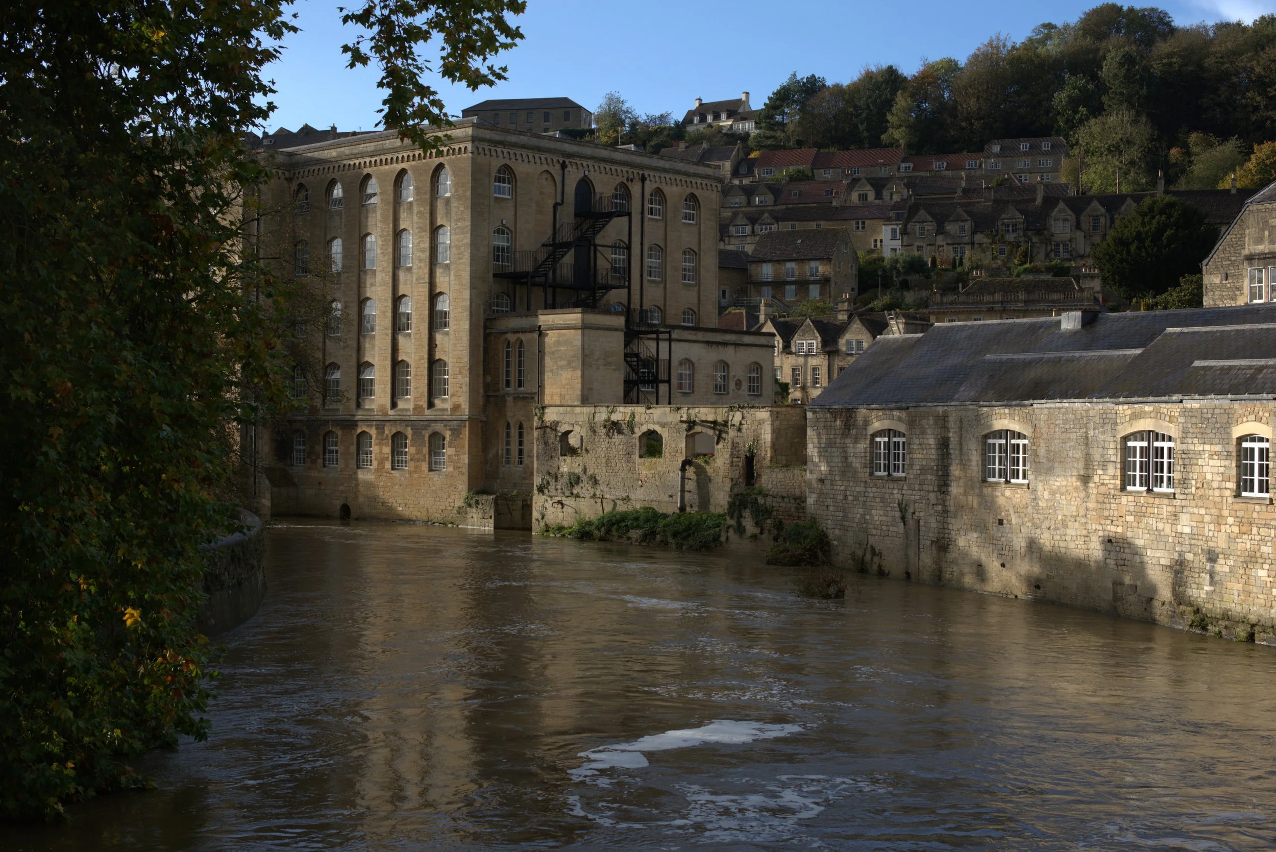 A river flows past old stone buildings with multiple windows, some with arched tops, under a hill with numerous houses. The sky is clear and blue.