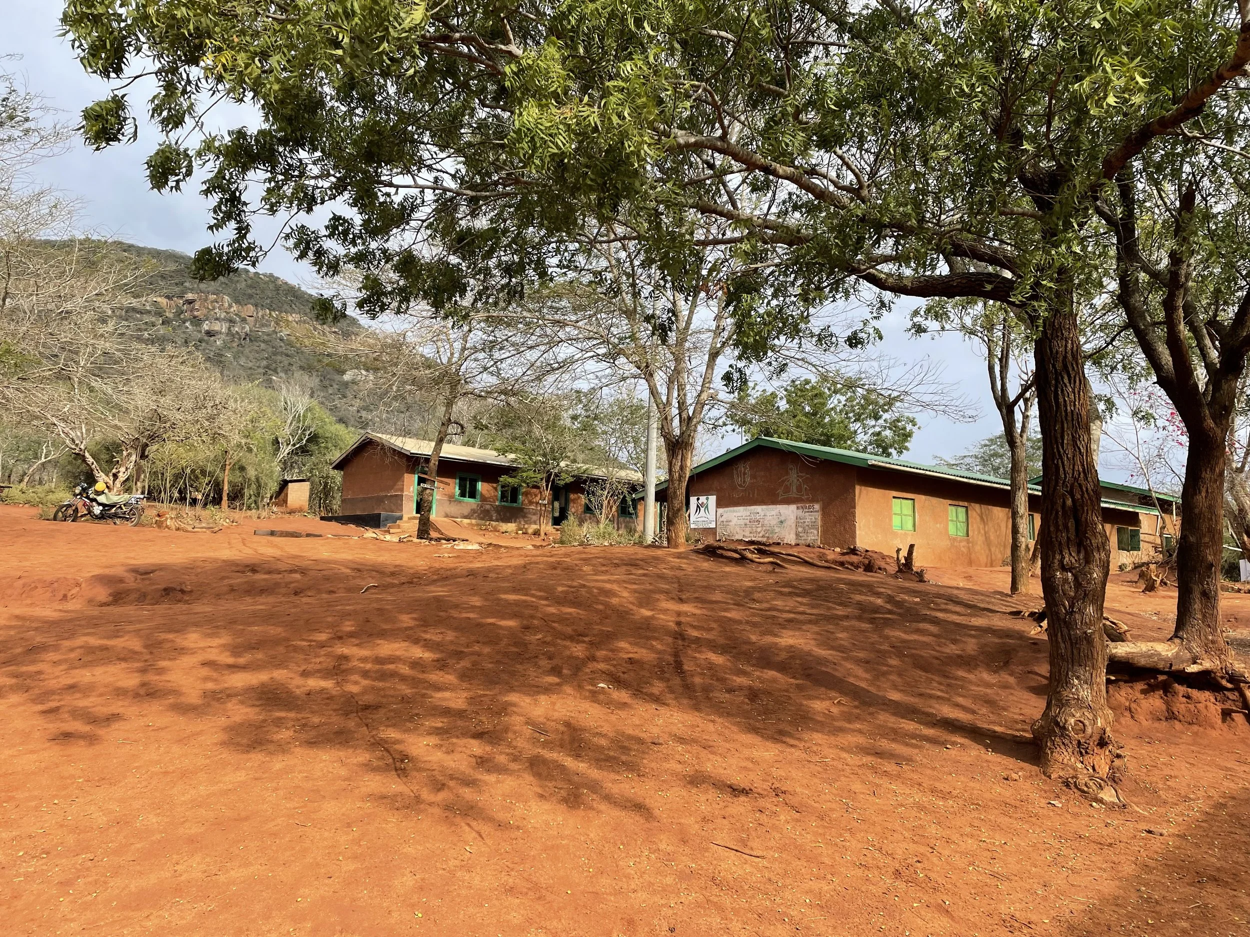 Rural scene with red dirt ground, scattered trees, two school buildings with green window frames, a motorcycle parked on the dirt, and hills in the background under a partly cloudy sky.