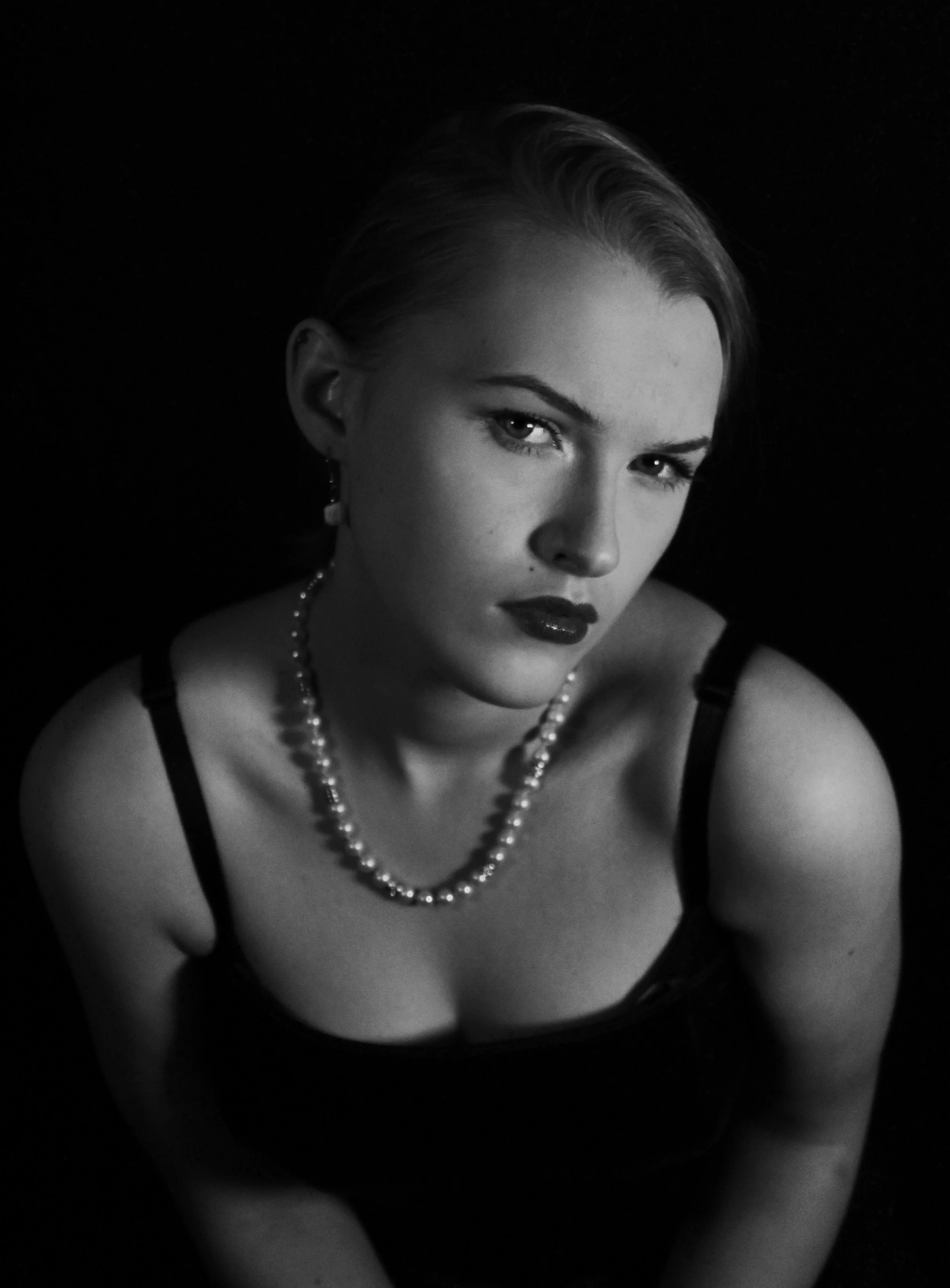 A black-and-white portrait of a woman with short hair, wearing a pearl necklace, earrings, and a black dress with spaghetti straps, looking directly at the camera.