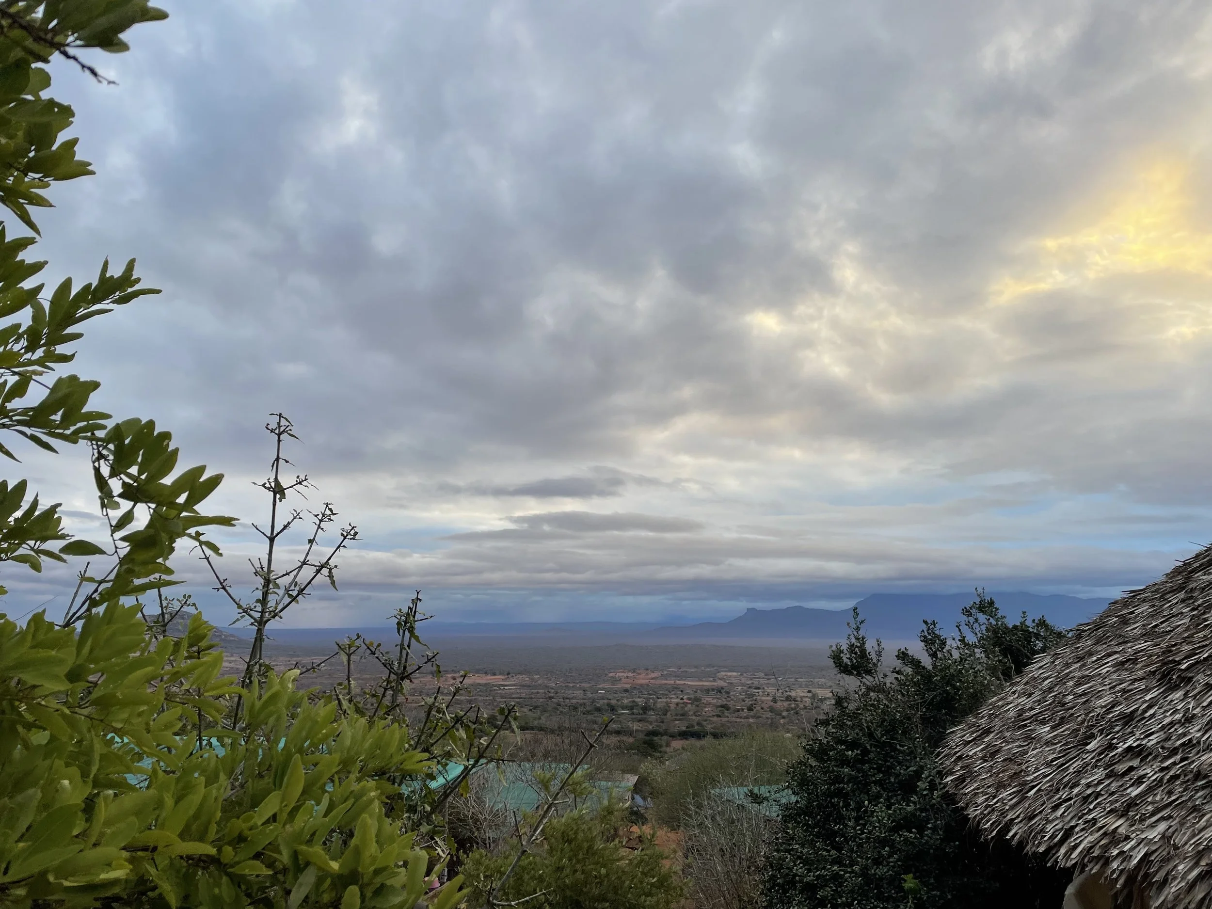 A landscape view in the evening of a vast plain with distant mountains, partially cloudy sky with hints of sunlight, and some greenery and thatched roof structures in the foreground.