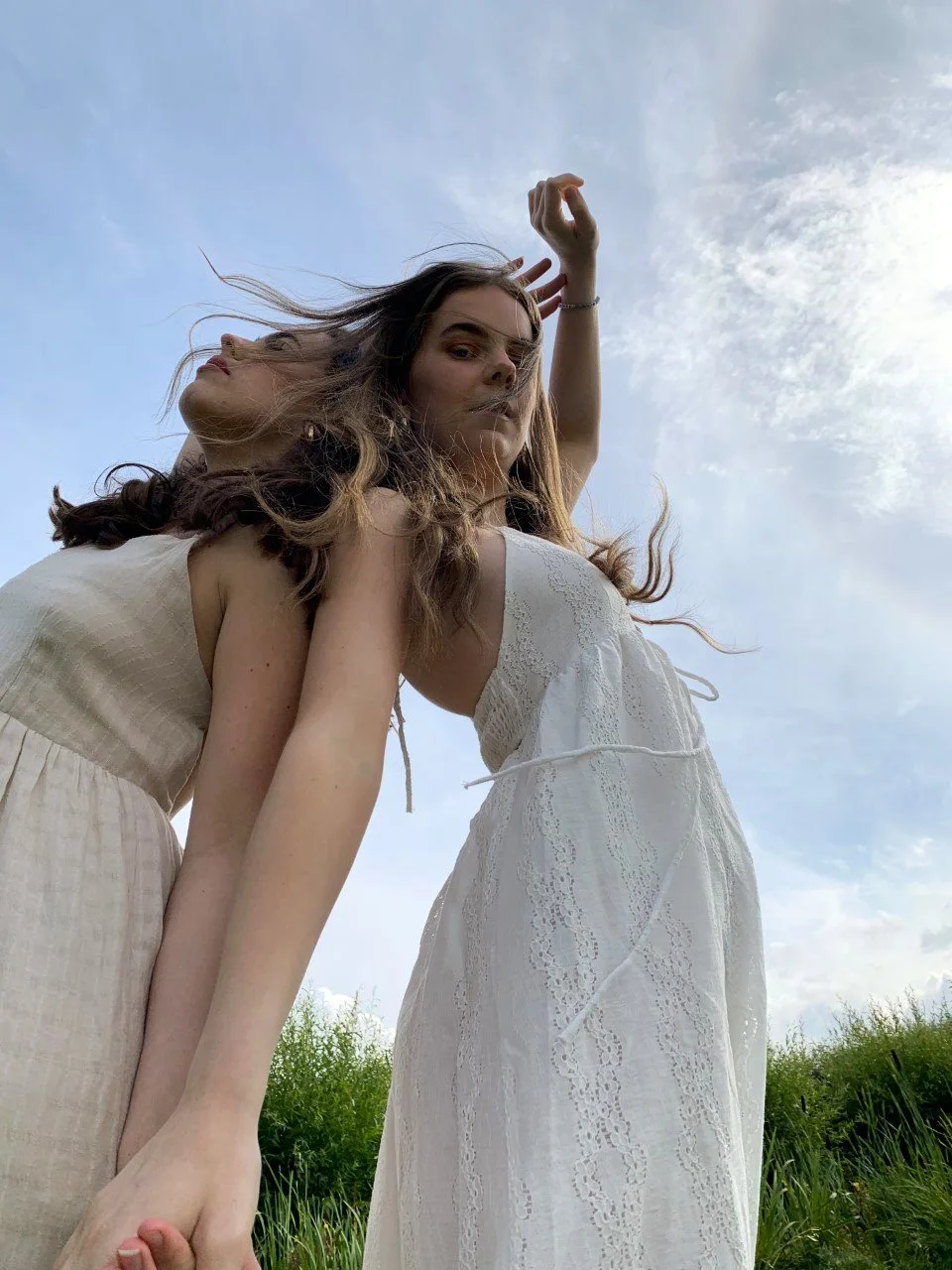 Two women in white dresses standing outdoors on a grassy field, seen from a low angle against a partly cloudy sky.