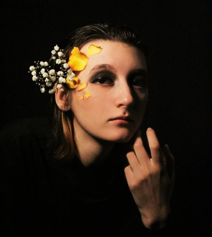 A woman with dark hair and light skin wearing dark makeup and earrings, with a floral hair accessory of yellow petals and small white flowers, posing against a black background.