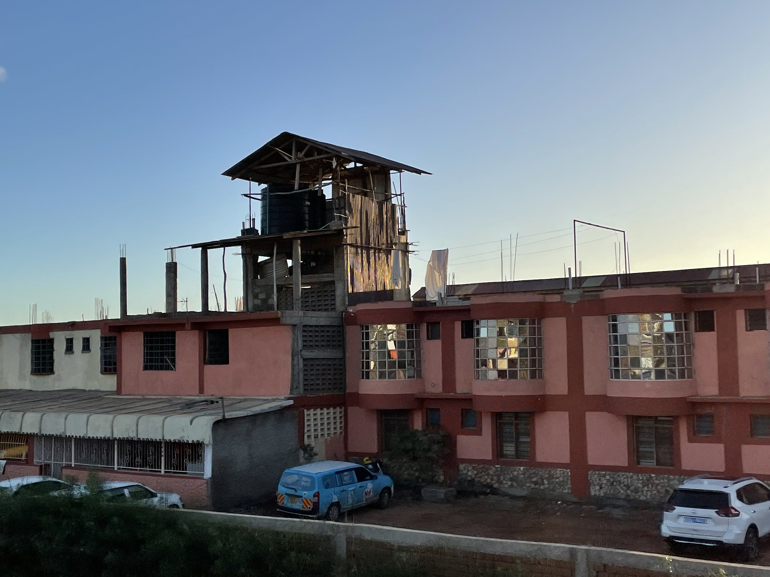 A pink building with glass block windows and a makeshift wood and metal structure on top, surrounded by parked cars and a sunset sky.