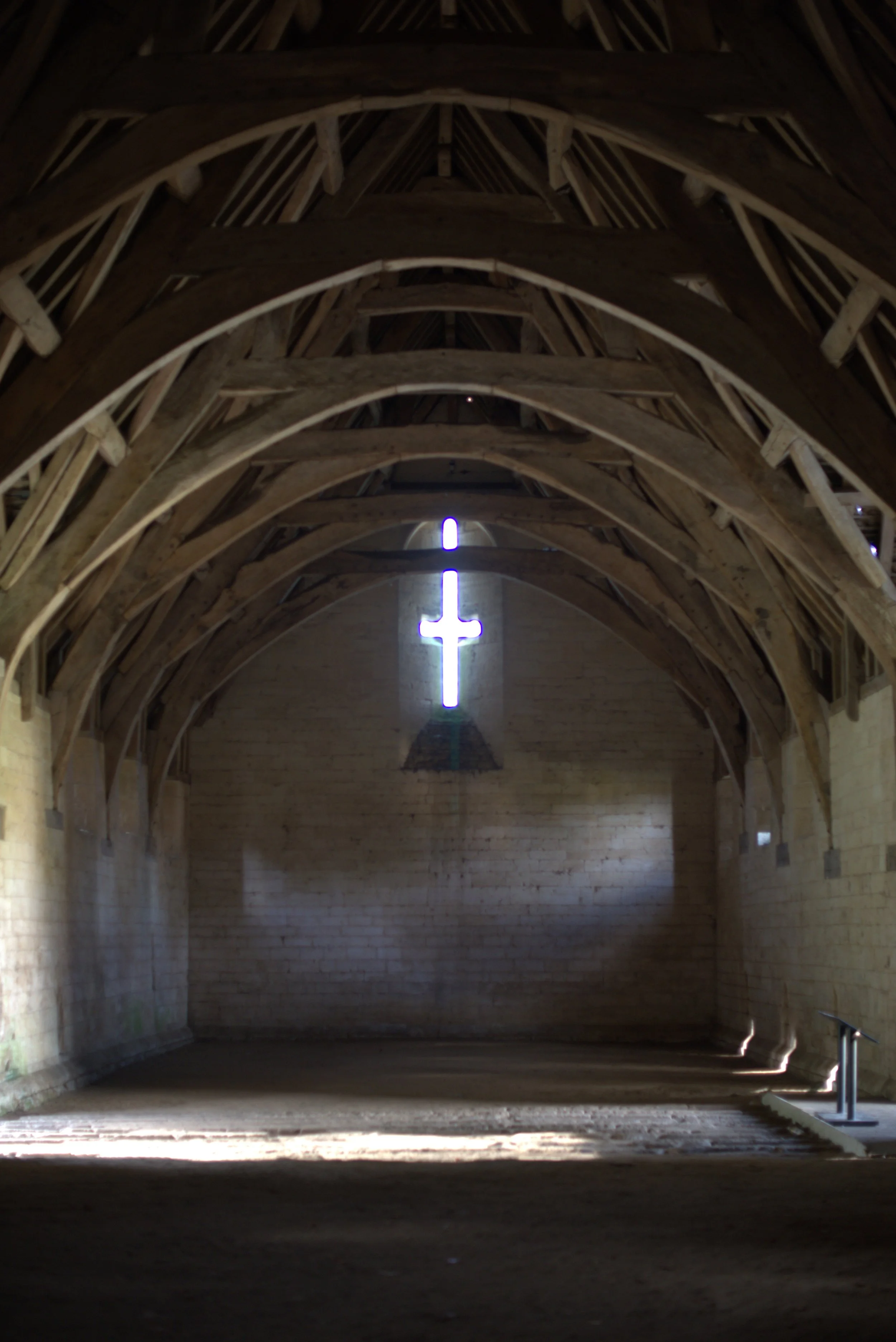 Interior of a church or chapel with a wooden vaulted ceiling, and a glowing cross-shaped opening in the wall allowing light to shine through.