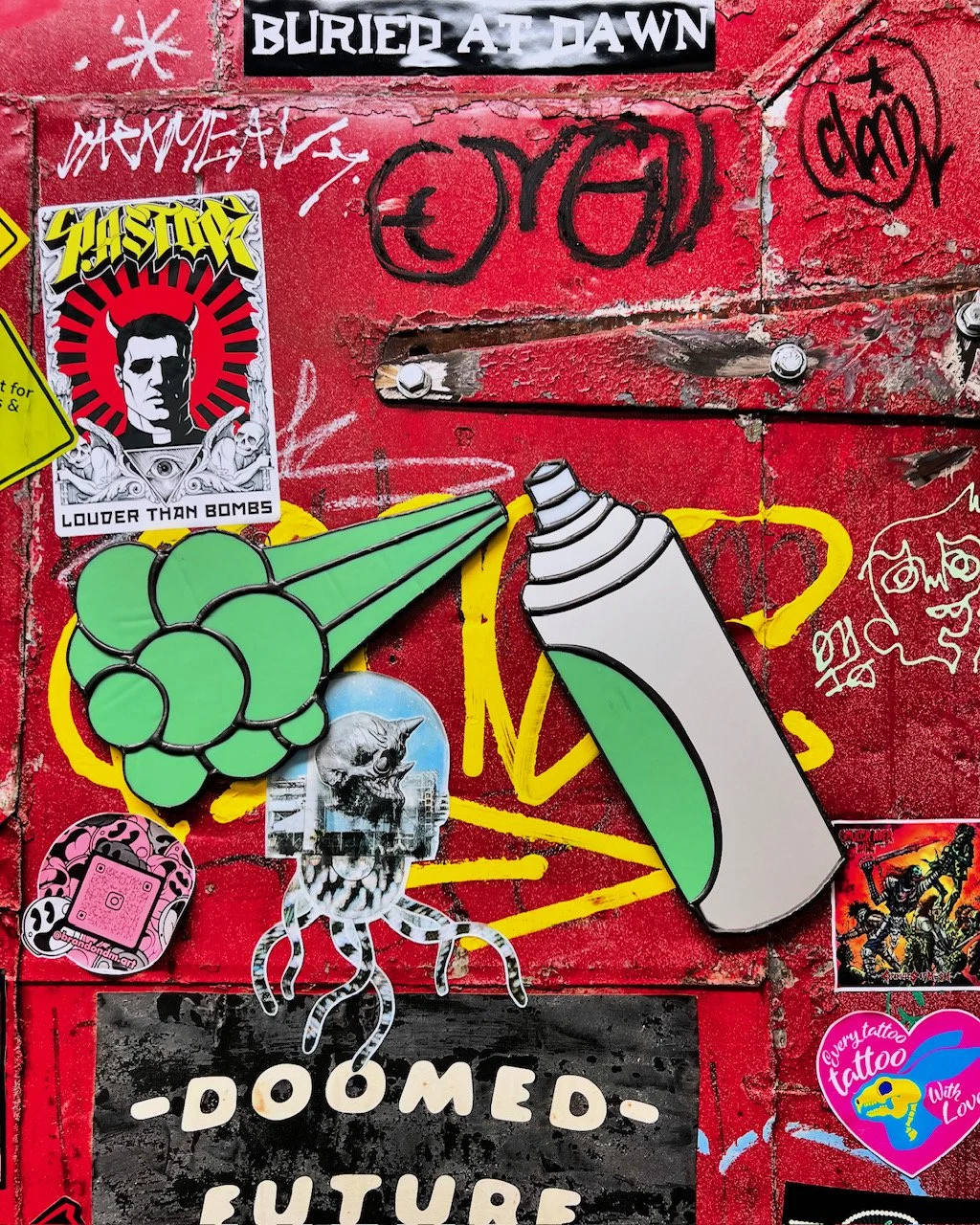 A graffiti-covered red wall decorated with various stickers and street art. One sticker features a black and white illustration of Frankenstein's monster with the phrase 'LOUder than bombs.' There are additional small stickers, including a pink Instagram logo, a cartoonish skull with tentacles, and a colorful tattoo sticker. The graffiti includes black and red tags and drawings, with a prominent yellow and green cartoon-style illustration of a bomb with a fuse.