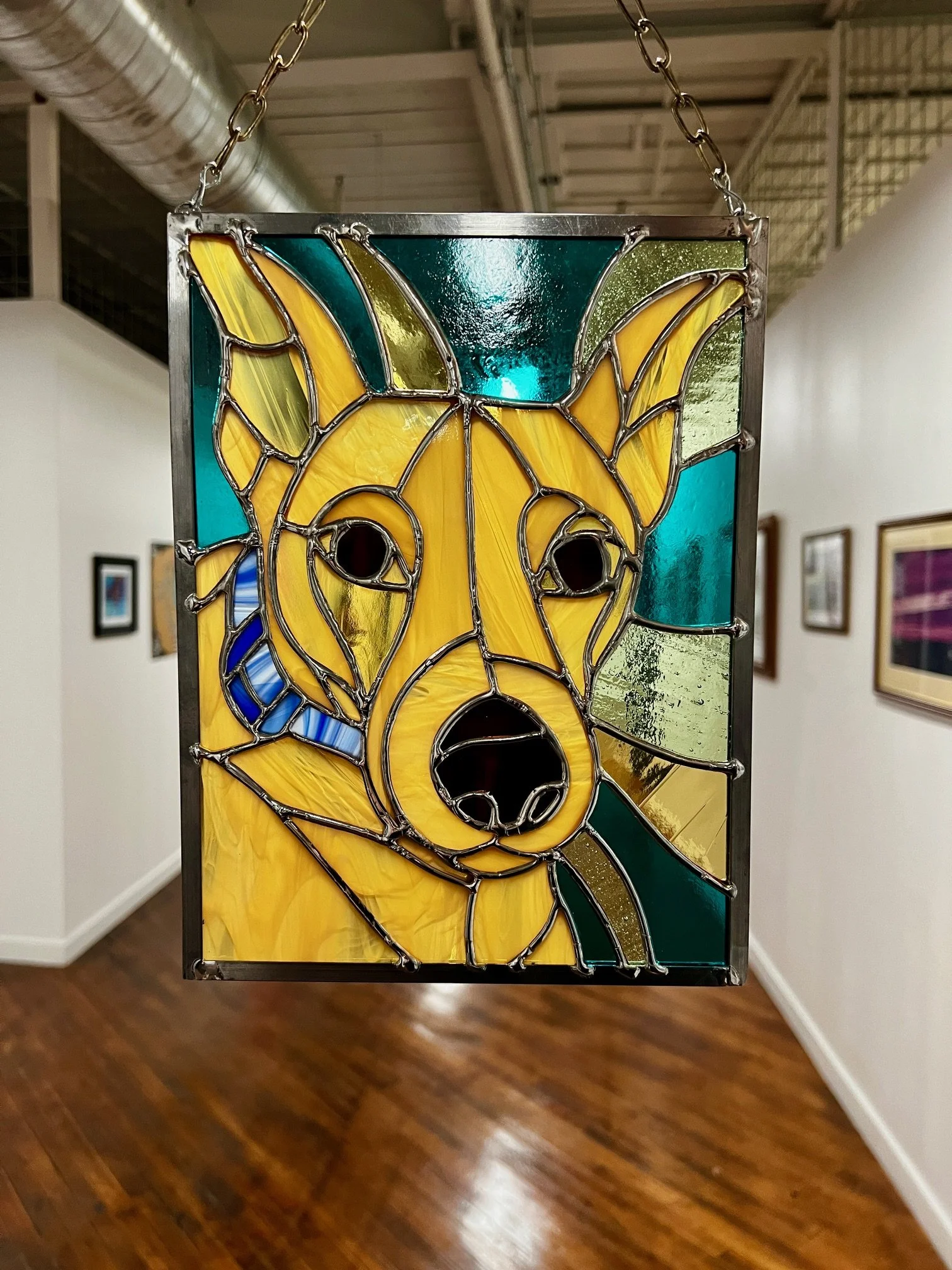 Colorful stained glass art piece depicting a dog's face with shades of yellow, green, and blue, hanging from a chain in an art gallery.