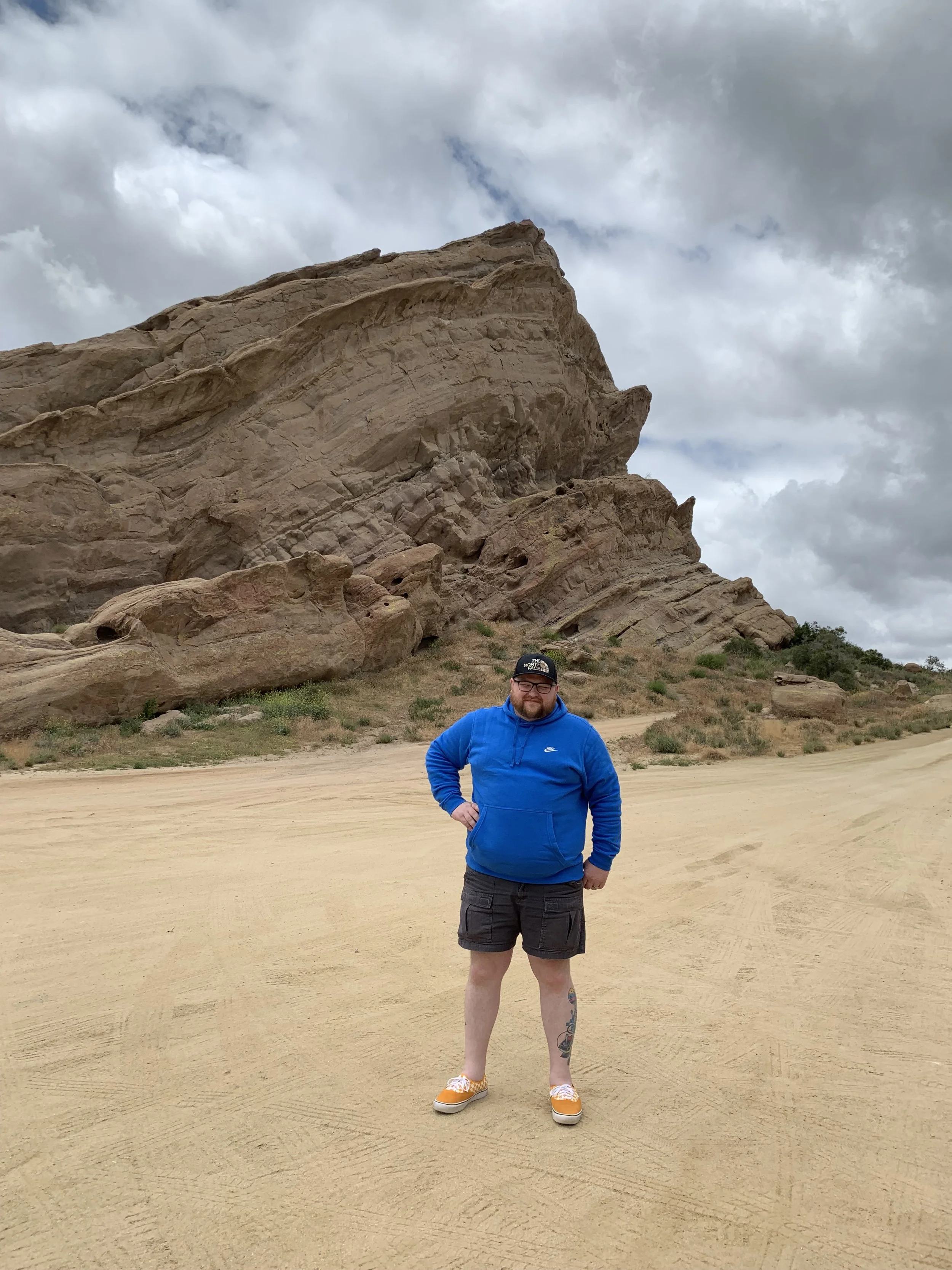 A man in a blue hoodie and shorts standing on sandy terrain with large rock formations and cloudy sky behind him.