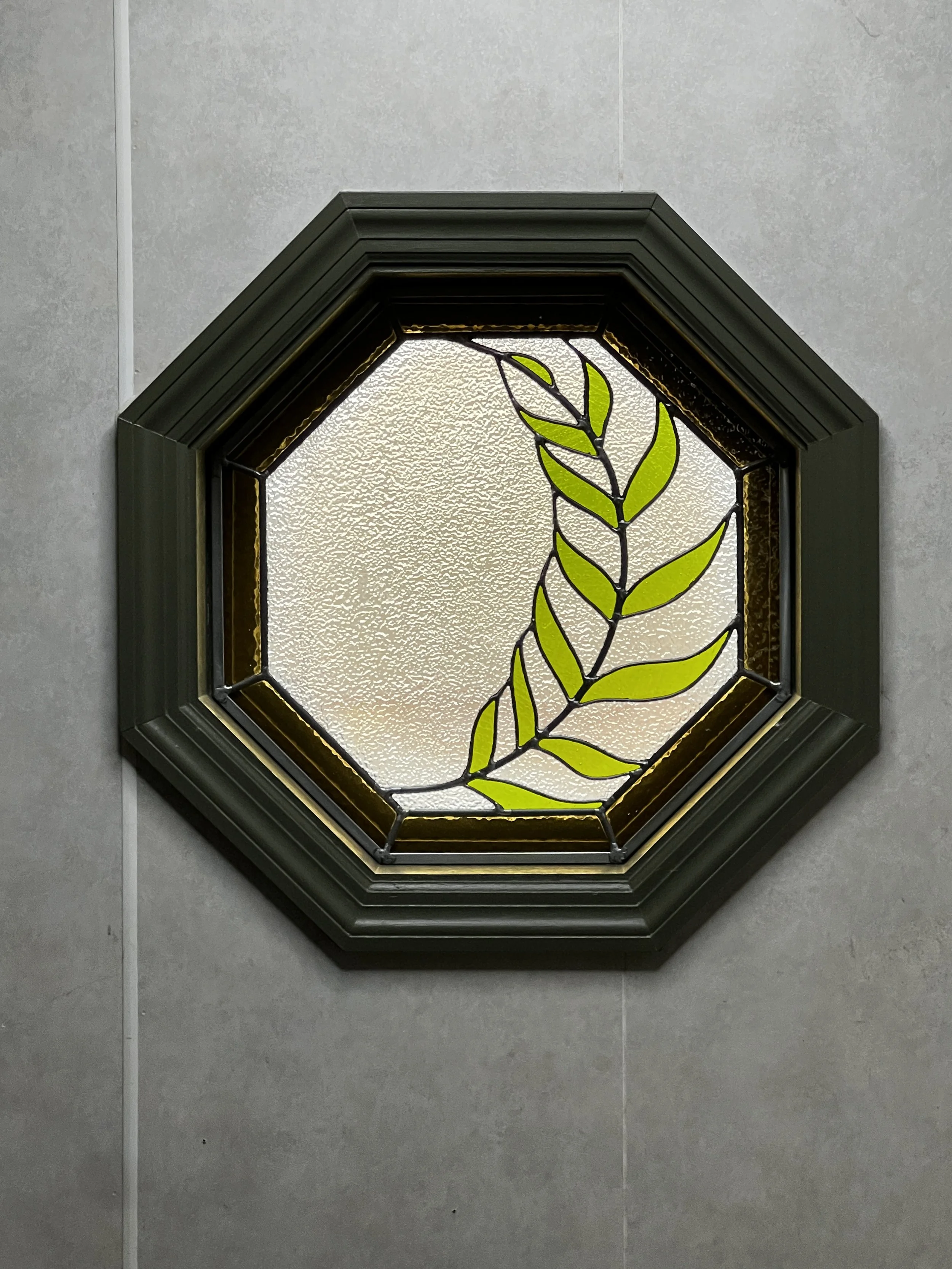 A stained glass window with an octagonal shape and a black frame, featuring a green leafy branch design on frosted glass.