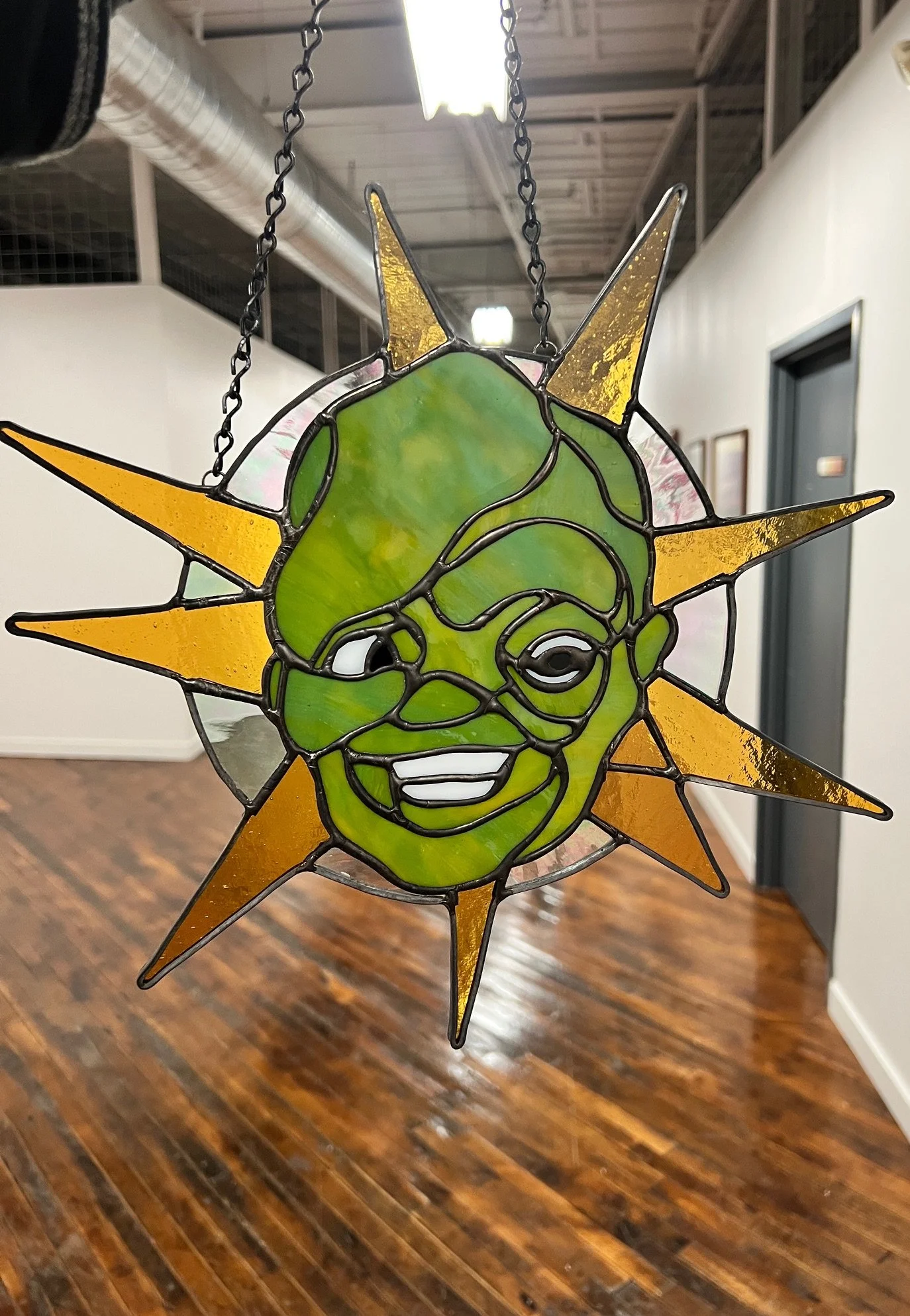 Stained glass art piece featuring a cheerful green alien face with smile and one eye, surrounded by orange and yellow sun rays, hanging from a chain.