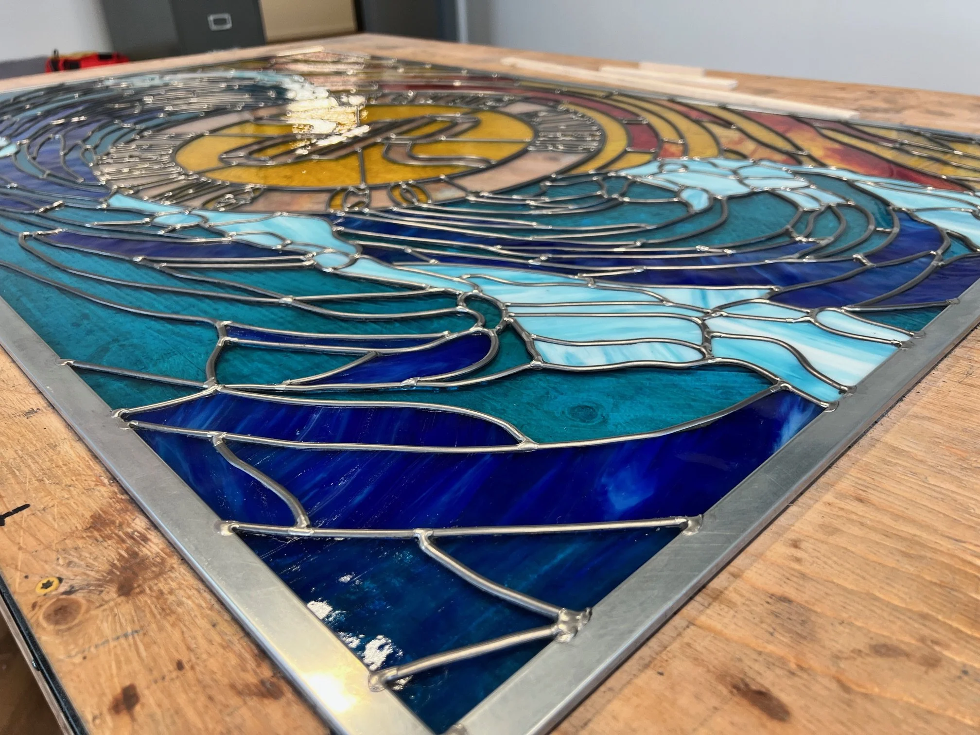 Close-up of a colorful stained glass artwork on a wooden table, featuring a swirling pattern with shades of blue, yellow, and red, held together by metal outlines.