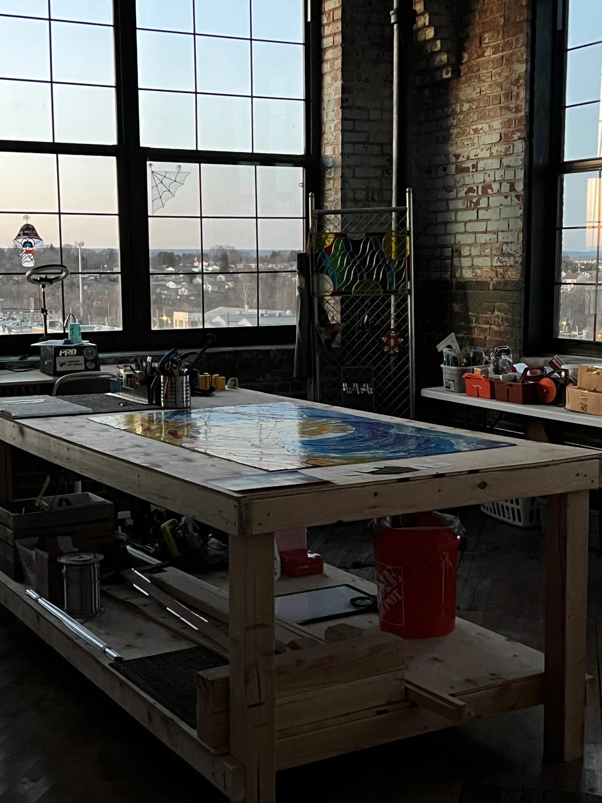 An artist's studio with a large wooden worktable, stained glass artwork, and tools. Large industrial windows reveal a cityscape outside.