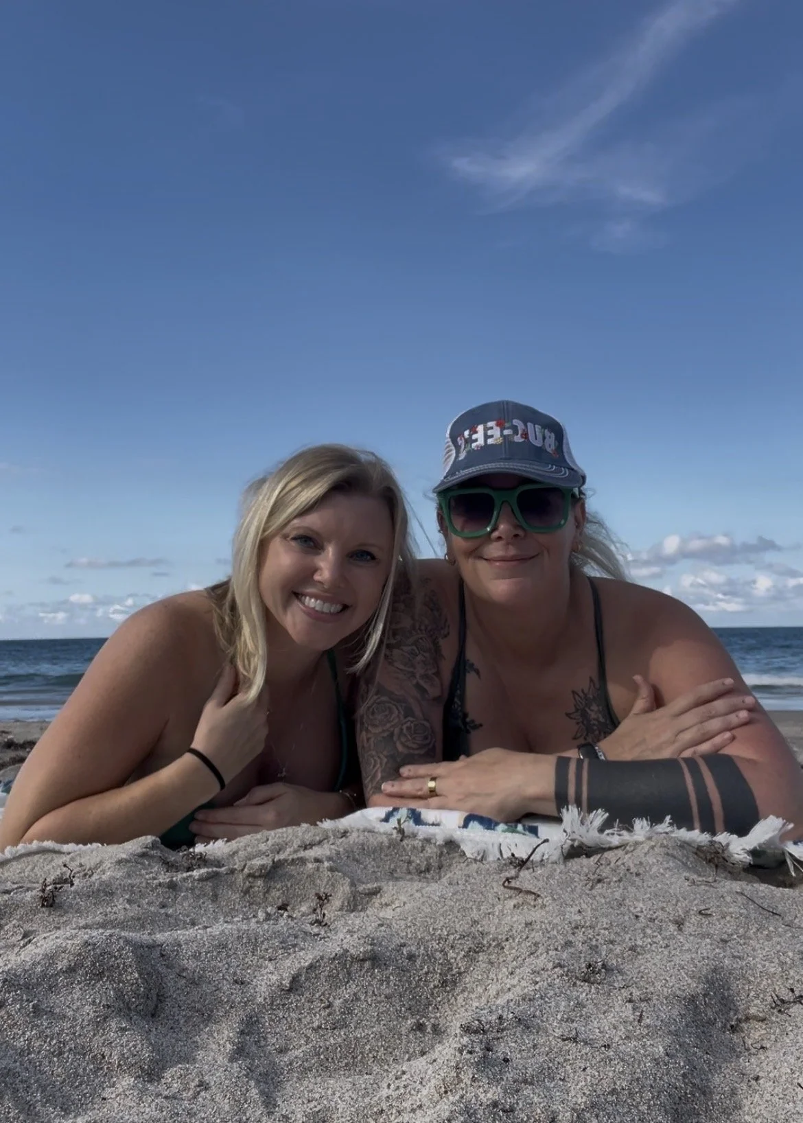 Two women lying on a sandy beach, smiling at the camera. The ocean and blue sky with some clouds are visible in the background.