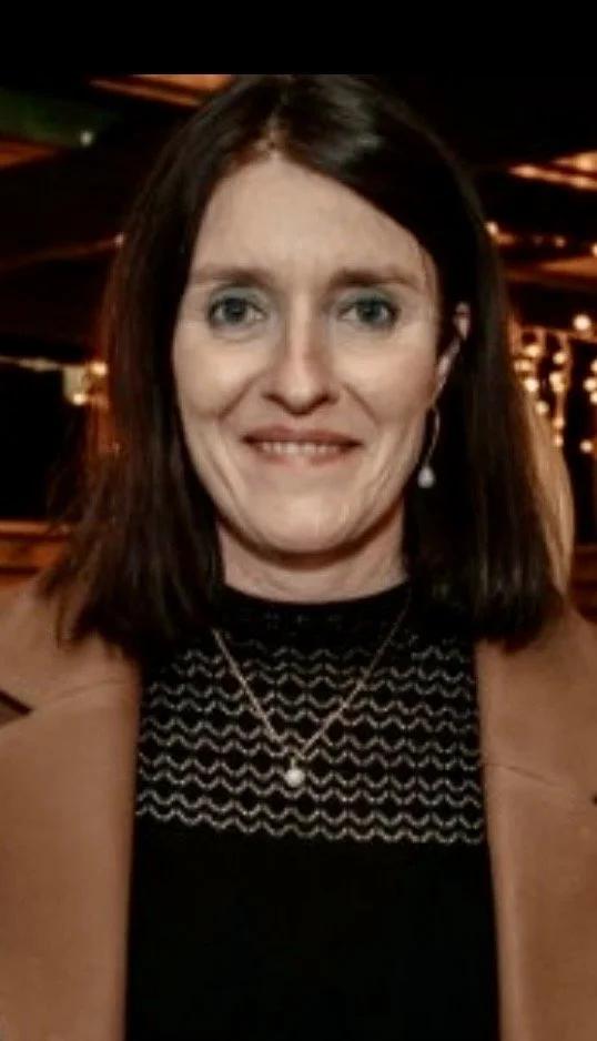 A woman with shoulder-length brown hair, wearing a black patterned top, light brown jacket, pearl necklace, and drop earrings, smiling at the camera in a dimly lit indoor setting with warm lighting and decorative lights in the background.