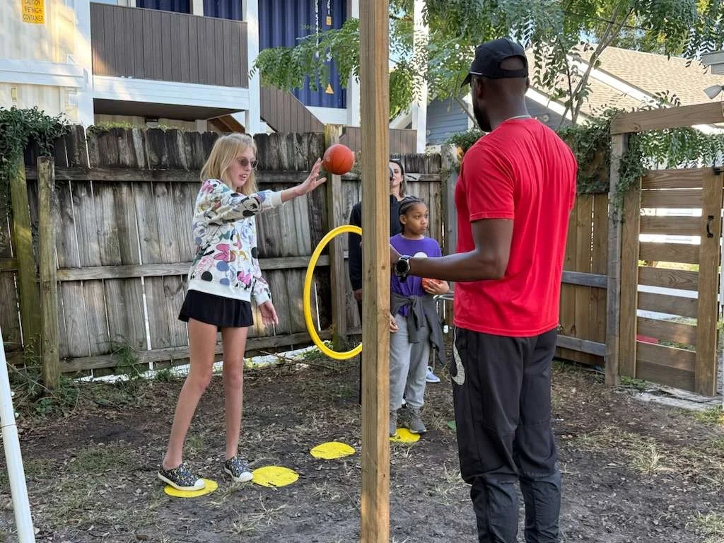 A young girl with blonde hair, wearing sunglasses, a colorful jacket, and a black skirt, is playing basketball on a backyard court. She is standing on yellow balance pads with circular footprints and is reaching out to catch or throw an orange basketball. An adult man wearing a red shirt and black cap is observing her, while two young girls, one with curly hair and the other with straight hair, watch from behind a wooden post on the fence.