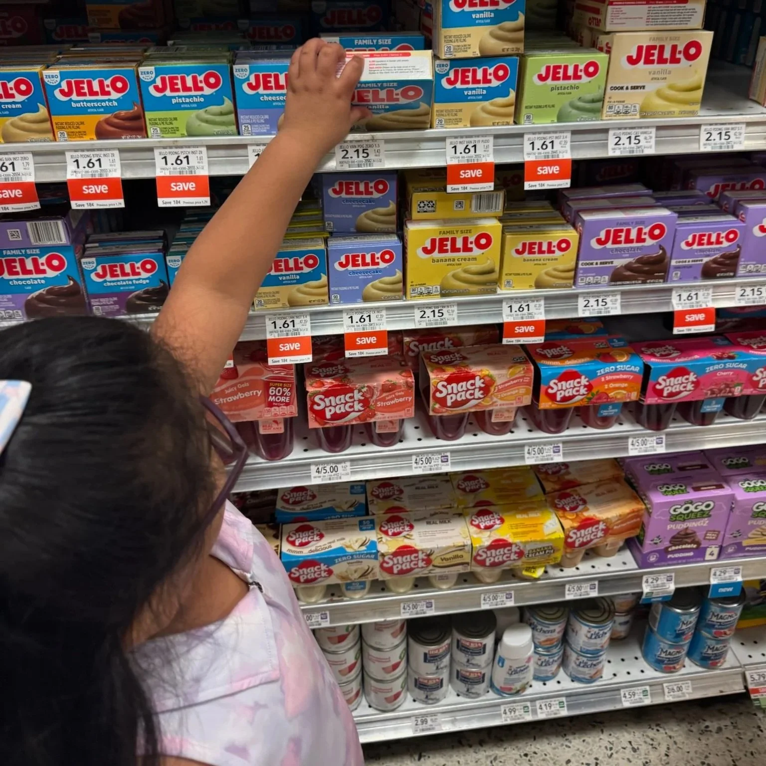 A young girl with glasses and a pink shirt reaching onto a supermarket shelf for a box of Jello vanilla pudding.