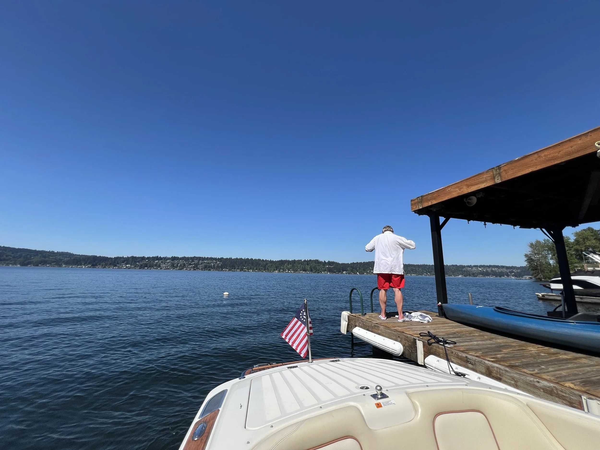 A man in red shorts and a white shirt standing on a wooden dock by a lake, with boats moored nearby and a clear blue sky.